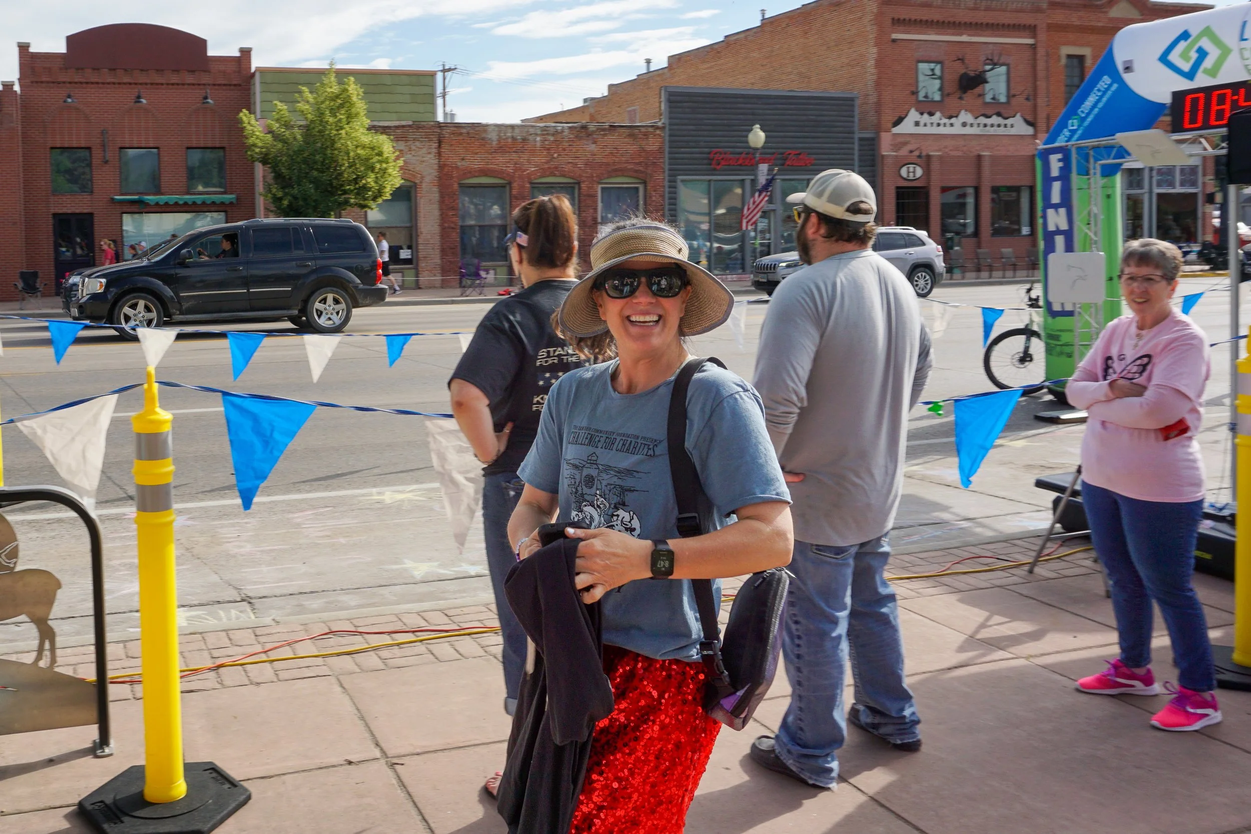 People gathered at a street event with banners and a timing clock, some are smiling and talking, brick buildings line the background under a partly cloudy sky.