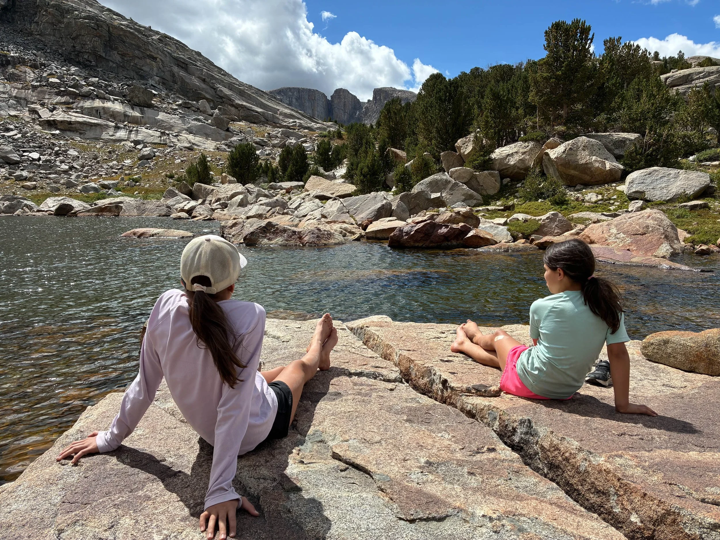 Two children sitting on rocks near a lake, with forested hills and mountains in the background under a partly cloudy sky.