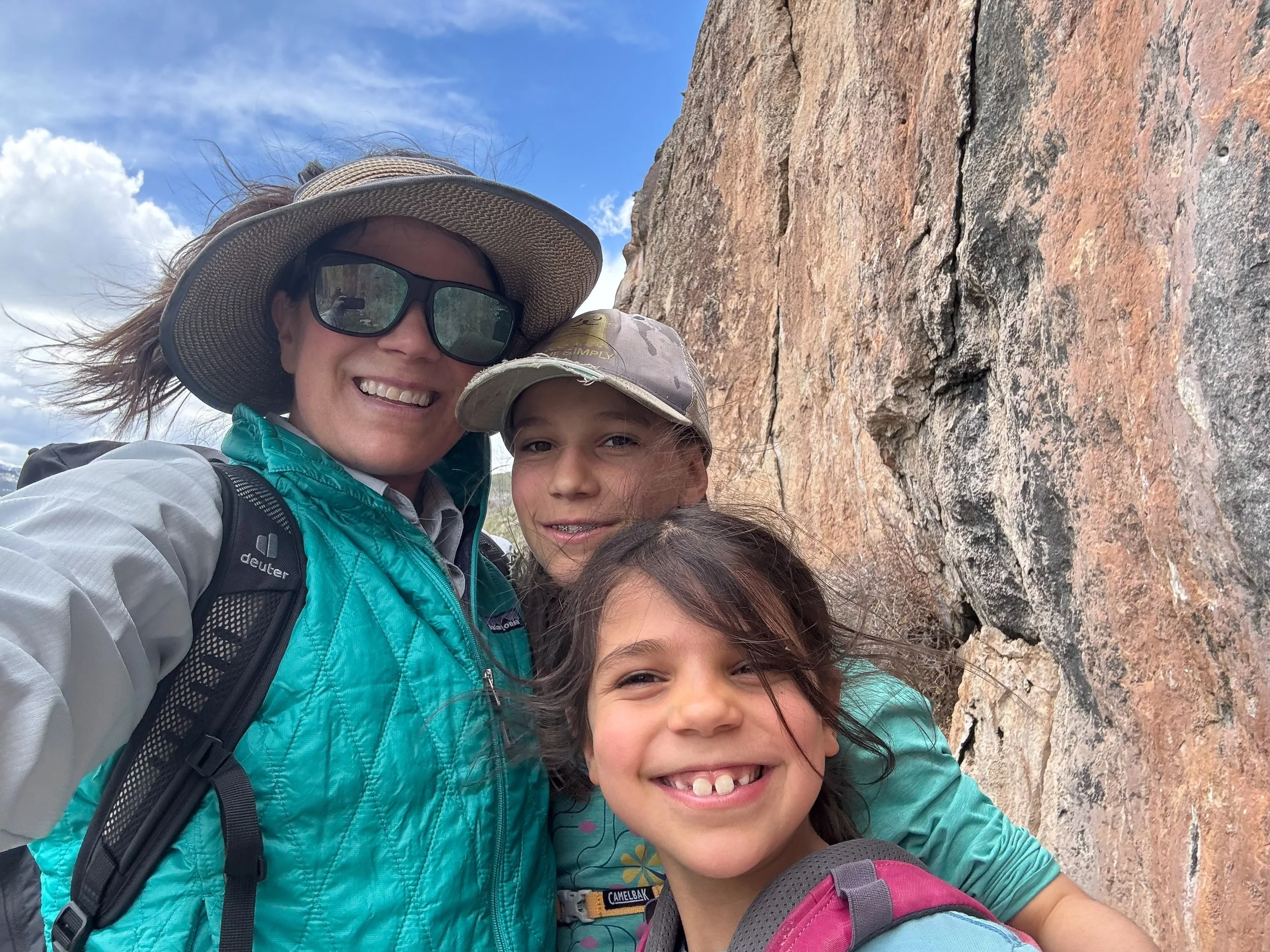 A woman with sunglasses and a wide-brimmed hat, and two children smiling, standing in front of a large rock formation outdoors during daytime.