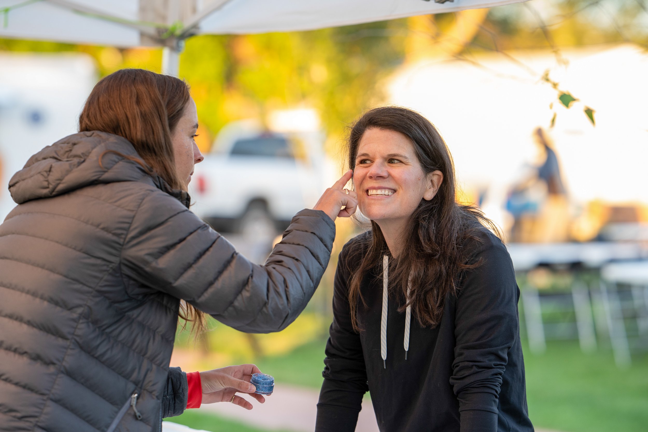 Two women having a conversation outdoors during daylight, one woman is applying makeup or powder to the other woman's face, with trees and a person walking in the background.