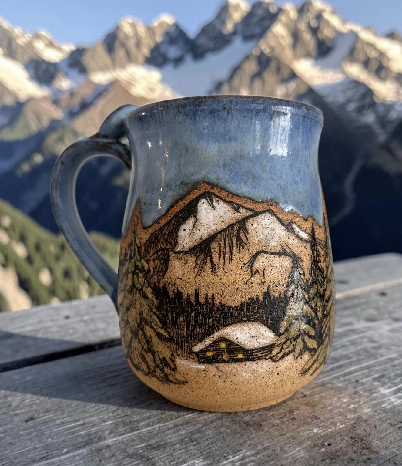 A ceramic mug with a mountain landscape painted on it, placed on a wooden surface outdoors with snow-capped mountains in the background.
