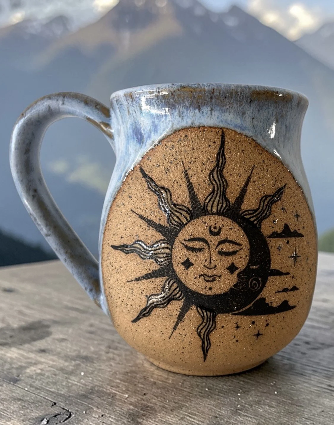 Ceramic mug with a cosmic sun and moon illustration, featuring a face on the moon, stars, and mountain silhouettes, placed on a wooden surface with mountains and sky in the background.