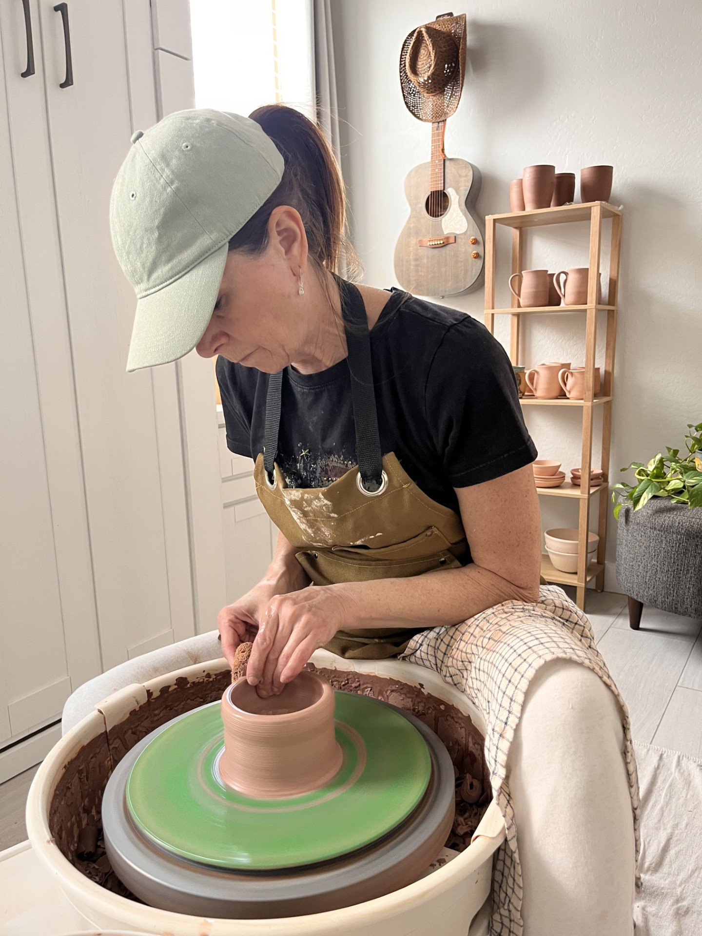 An elderly woman working on a pottery wheel shaping clay into a cylinder in a cozy room decorated with a guitar, hat, and shelves of pottery.
