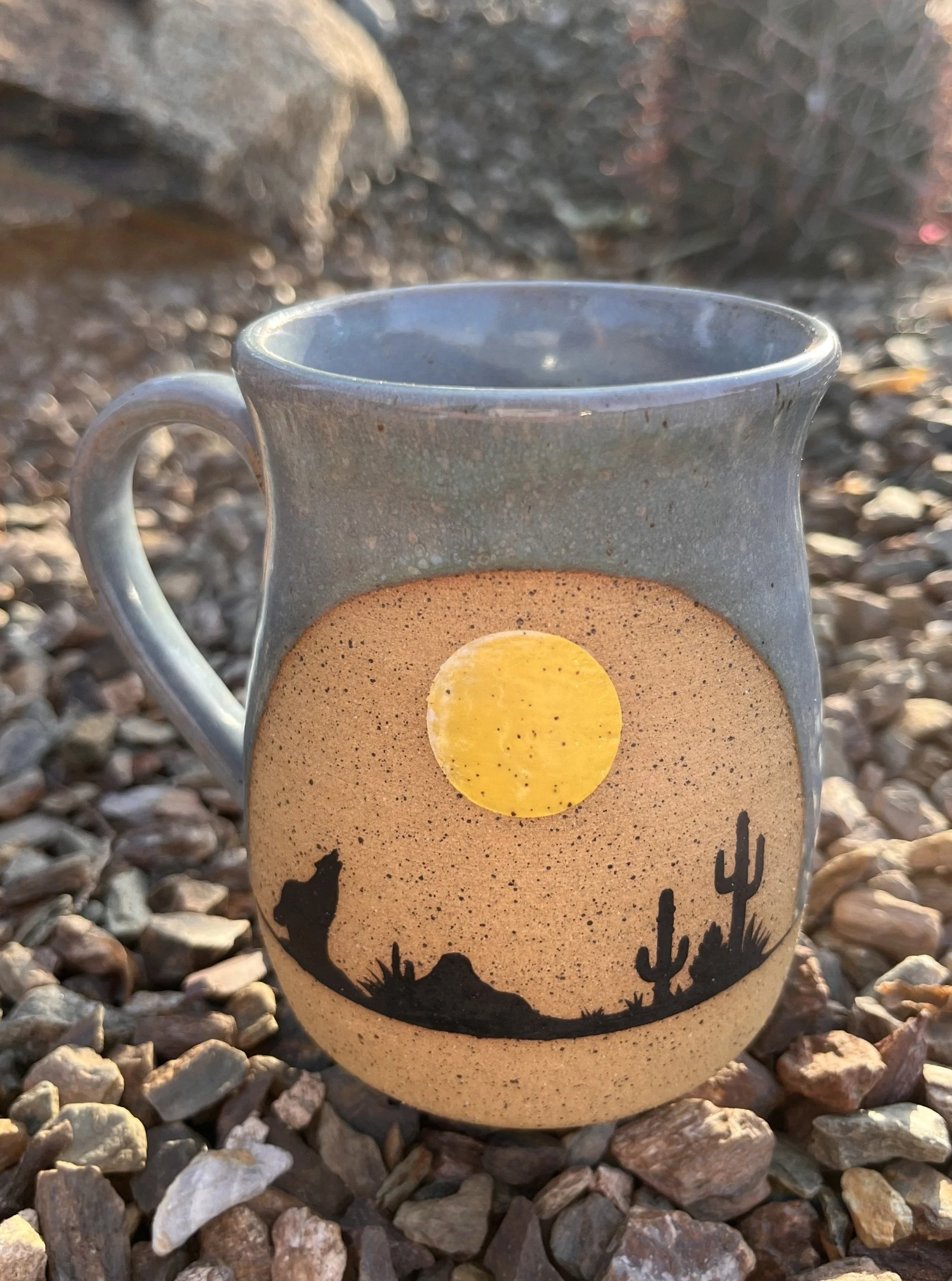 A ceramic mug with a desert scene silhouette design, featuring cacti and a mountain range, placed on a bed of small rocks.