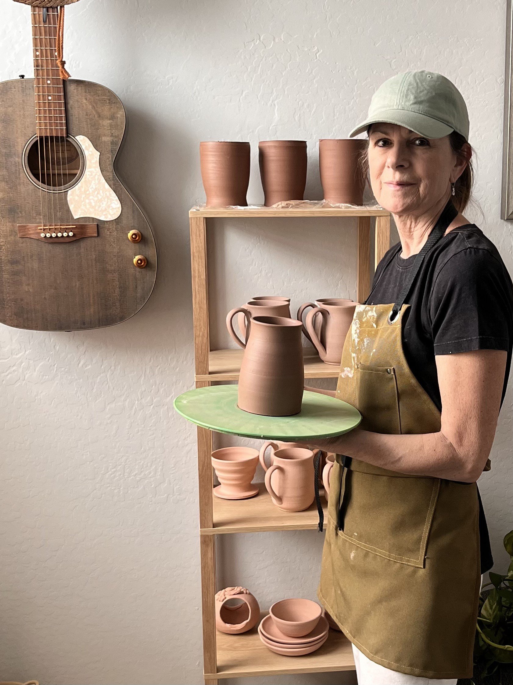 A woman in a beige apron and cap stands in a ceramics studio holding a round green tray with a large unfinished clay vase on it. Behind her, there is a wooden shelf with various unfinished clay pottery pieces such as mugs, bowls, and vases, and a wall-mounted wooden guitar.