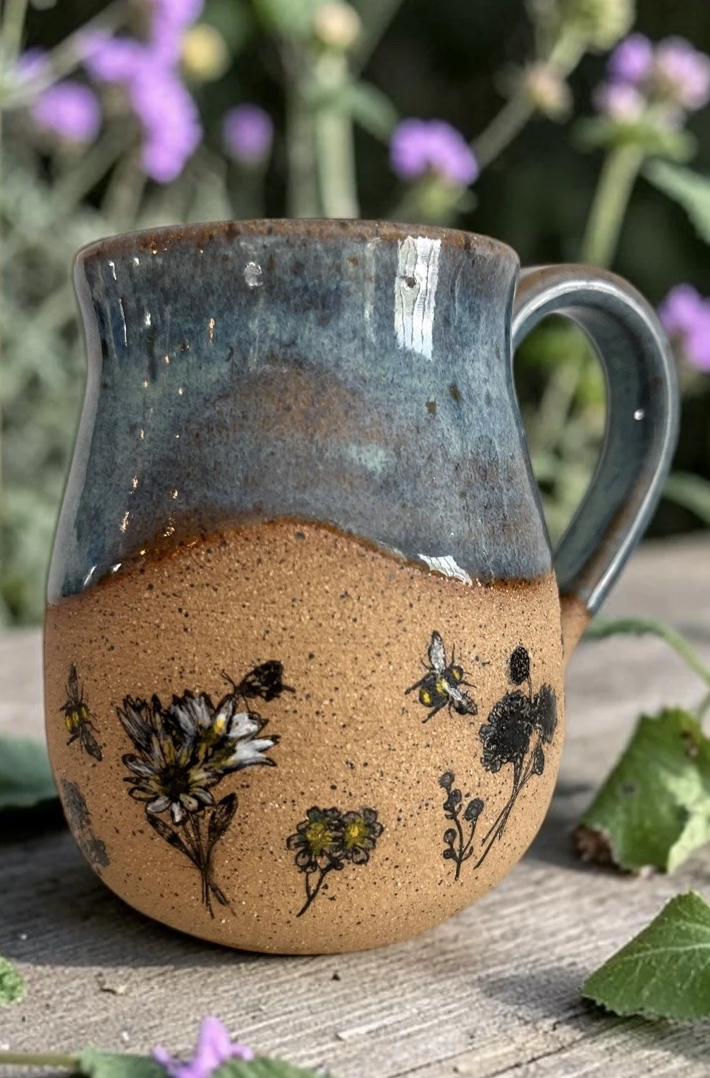 A ceramic mug with a blue and brown glaze, decorated with black and yellow bee and flower illustrations, placed on a wooden surface with green leaves and purple flowers in the background.