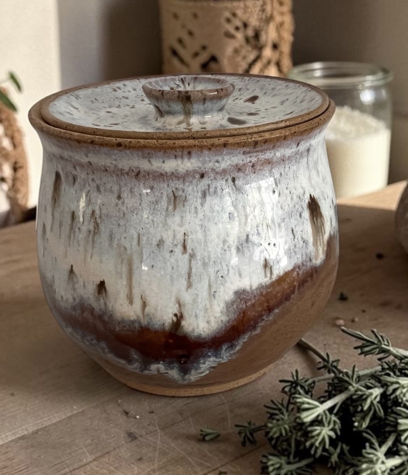 A rustic ceramic jar with a lid, glazed in a white and brown earthy tone, placed on a wooden surface with sprigs of greenery nearby.