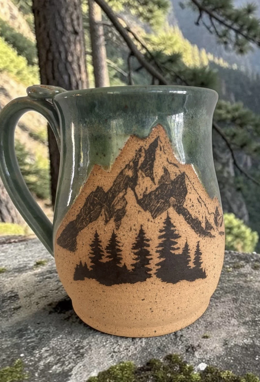 A ceramic mug with a mountain and forest scene painted on it, set outdoors on a rock with trees and mountains in the background.