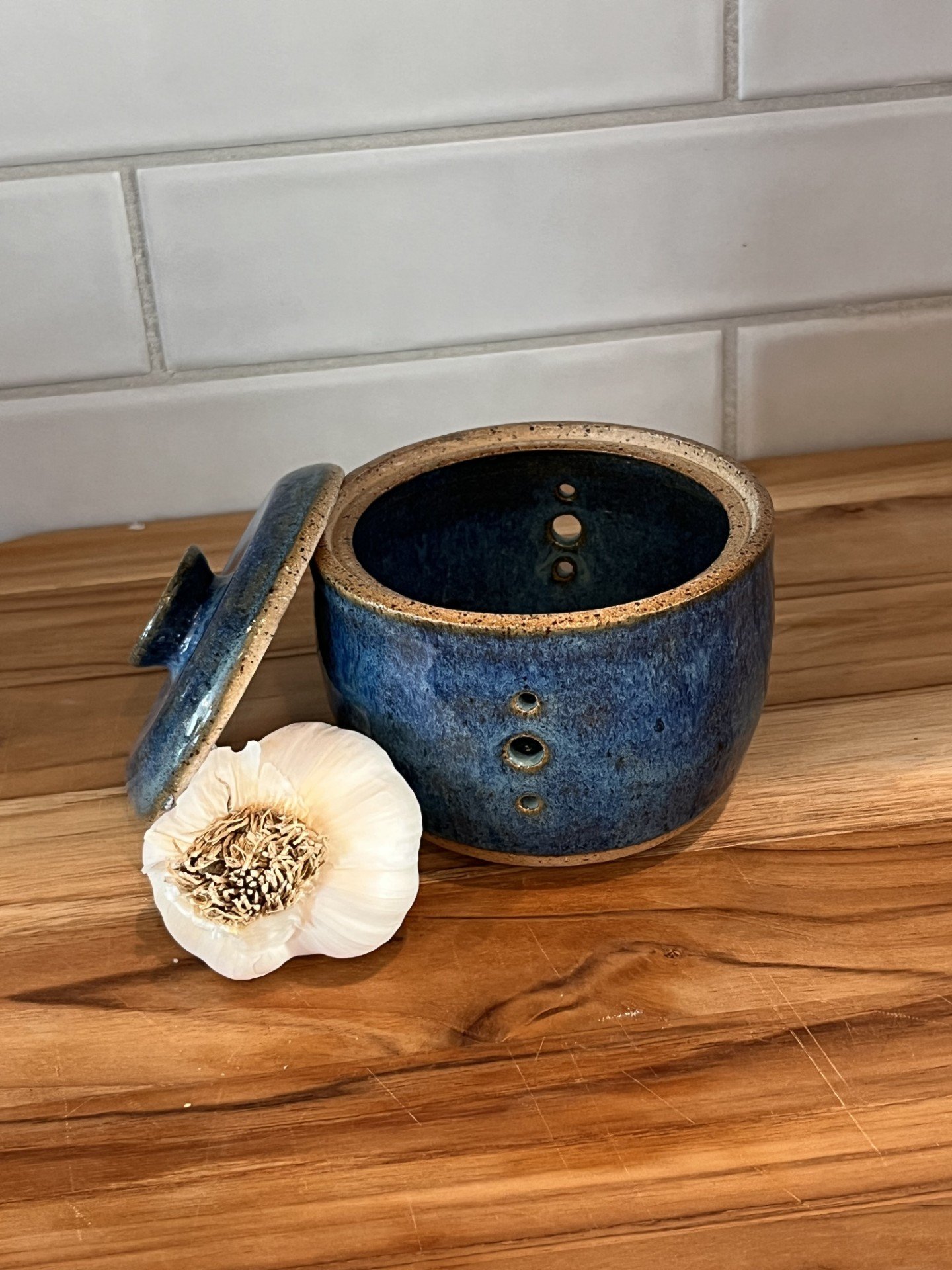 A blue ceramic pot with a lid placed to the side, on a wooden surface, with a white garlic bulb with flaky skin and a dried flower nearby, and a white tiled wall in the background.
