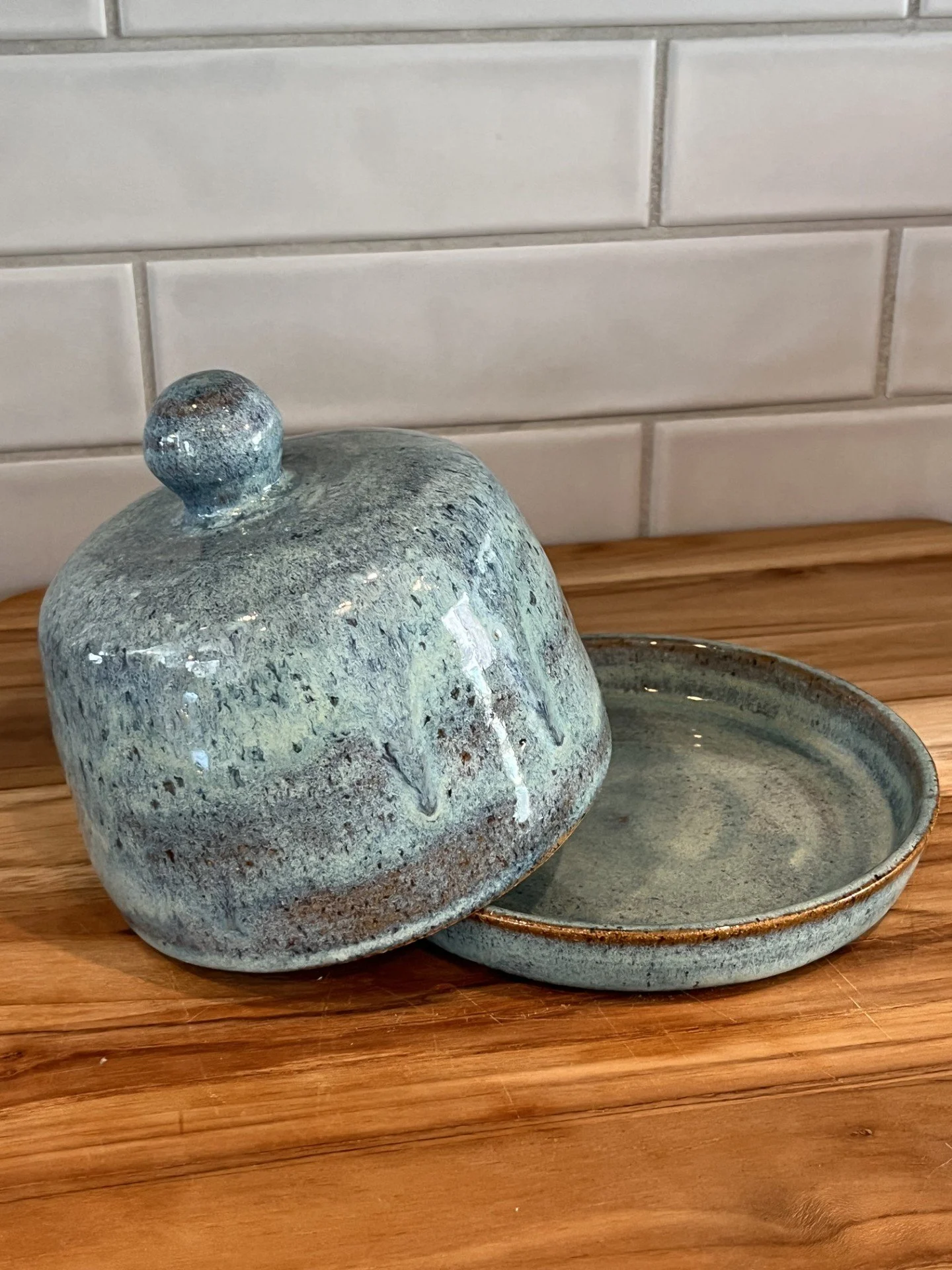 A ceramic stoneware butter keeper with a terraced lid next to a matching shallow dish, placed on a wooden surface with a white tile wall background.