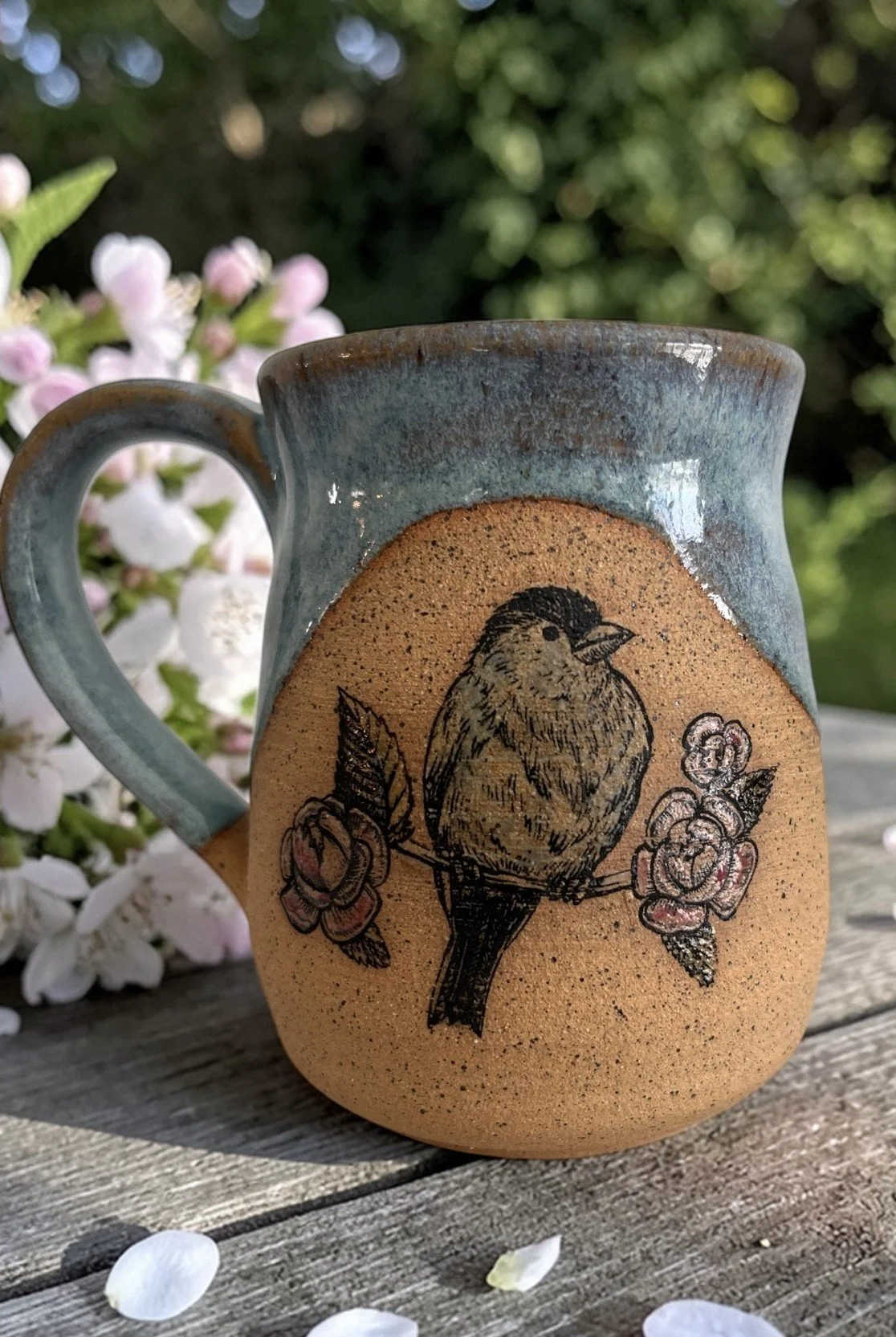 A ceramic mug with a painted bird perched on a branch surrounded by flowers. The mug is placed on a wooden surface with white flower petals scattered around and greenery in the background.
