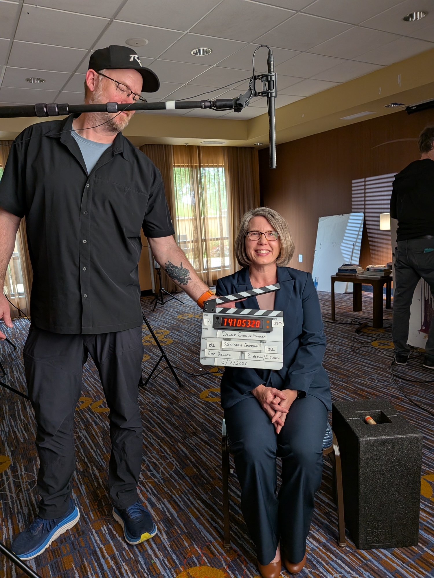 Dr. Karie Gibson sitting in a chair &  smiling, A man with glasses & black cap,  standing next to her, holding a clapperboard with cast and scene details adjusting a microphone overhead d in a room with wood paneling, windows, and a table with books.