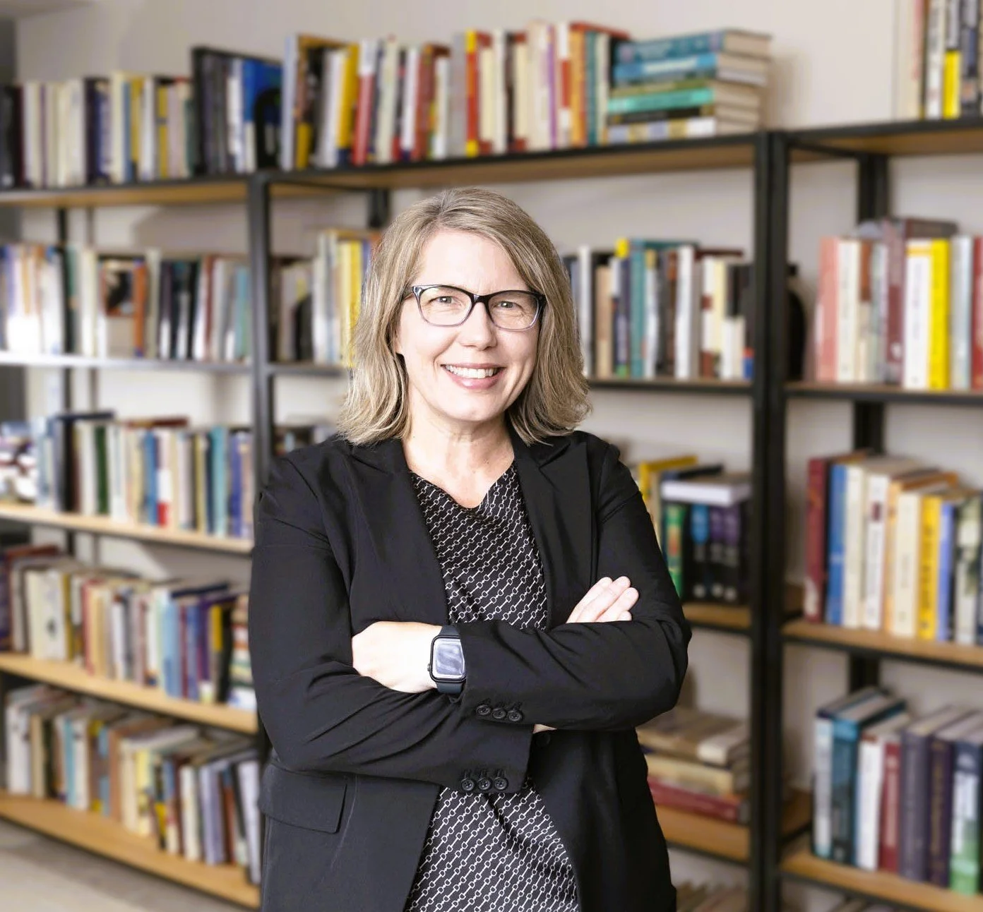 Dr. Karie Gibson. She is a woman with glasses and shoulder-length blonde hair, smiling with arms crossed, standing in front of bookshelves filled with books.