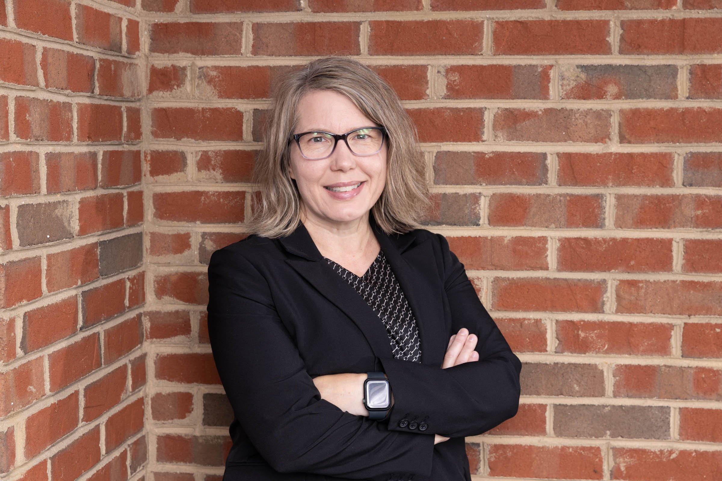 Dr. Karie Gibson. She is a woman with blonde hair, glasses, and a black blazer standing against a brick wall, smiling with arms crossed.