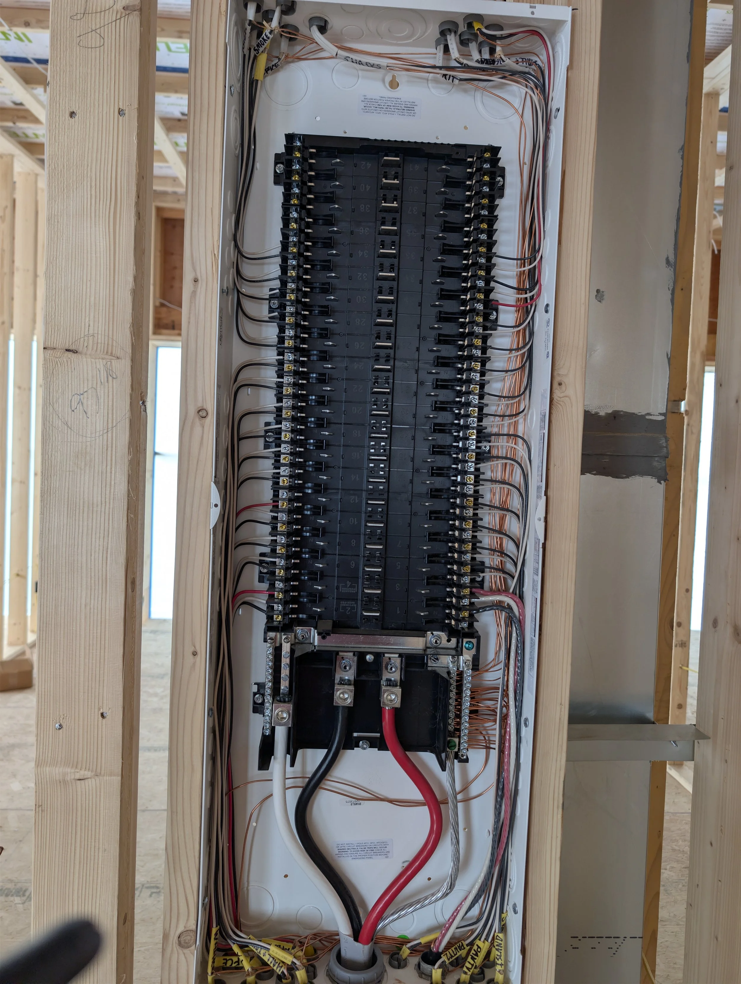 Interior view of an electrical breaker panel installed in a wood framing wall at a construction site, showing multiple circuit breakers and wiring. Clean organized wiring