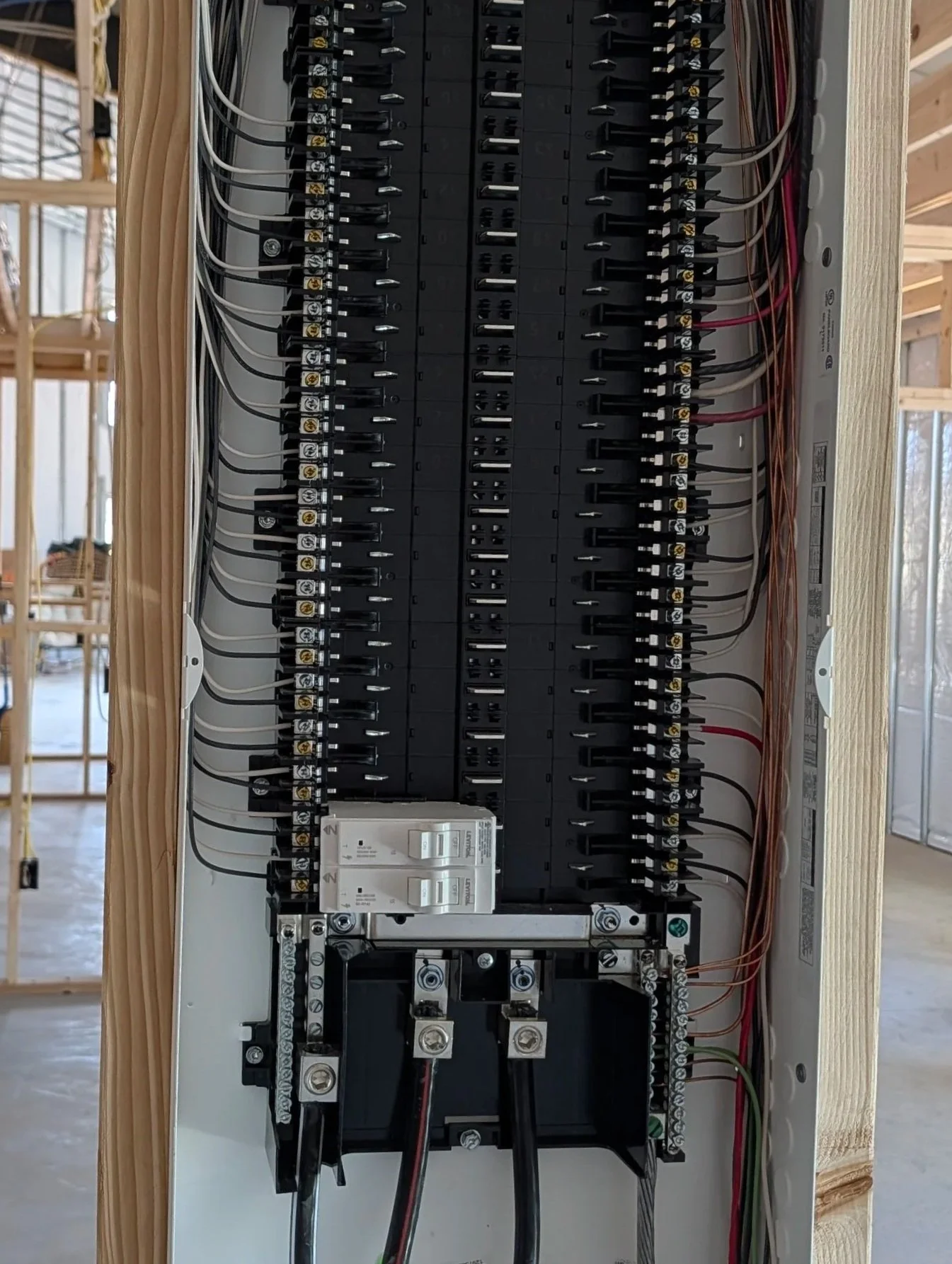Electrical panel with circuit breakers and wiring inside a wooden frame at a construction site.