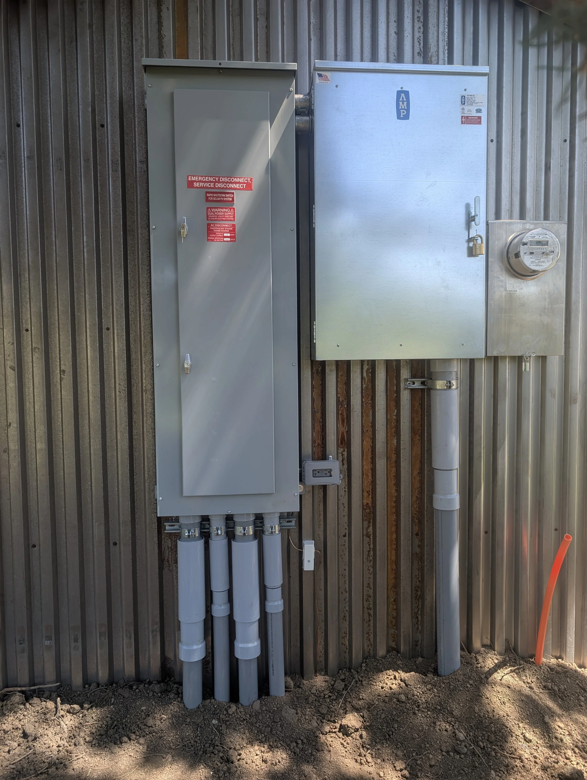 Electrical and solar panel utility boxes mounted on a corrugated metal wall, with pipes and an electrical outlet below, on dirt ground.