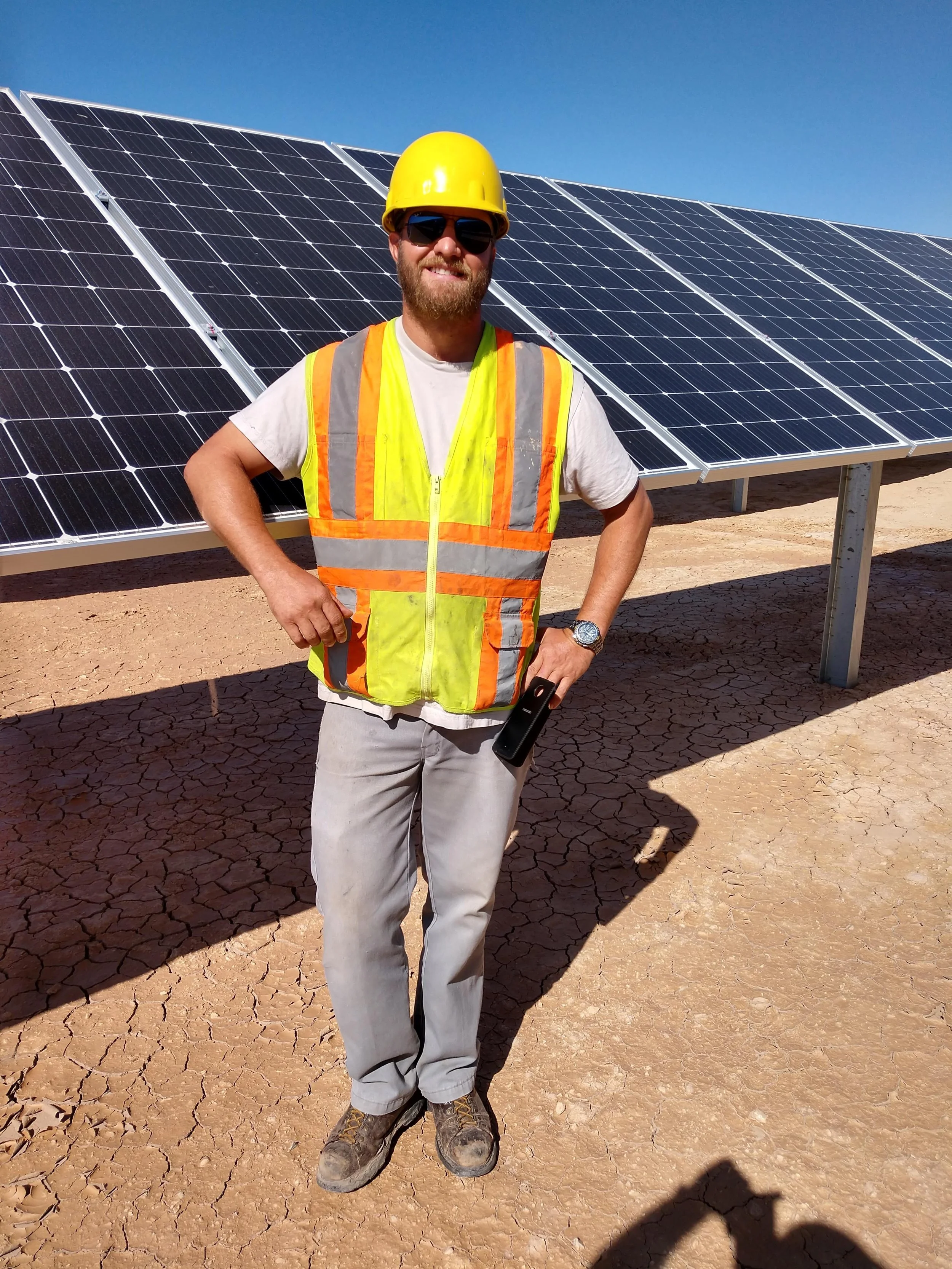 A man wearing a yellow safety helmet, sunglasses, a bright orange and yellow safety vest, a watch, a gray t-shirt, and gray pants stands in front of solar panels on a dry, cracked ground under a clear blue sky.