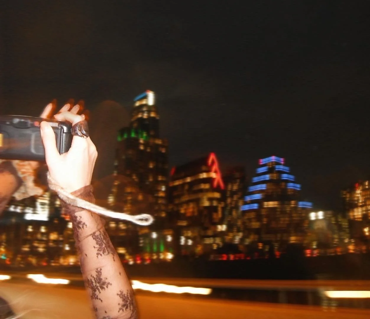 City skyline at night with illuminated skyscrapers and a person taking a photo with a phone, wearing lace gloves with a snowflake pattern.