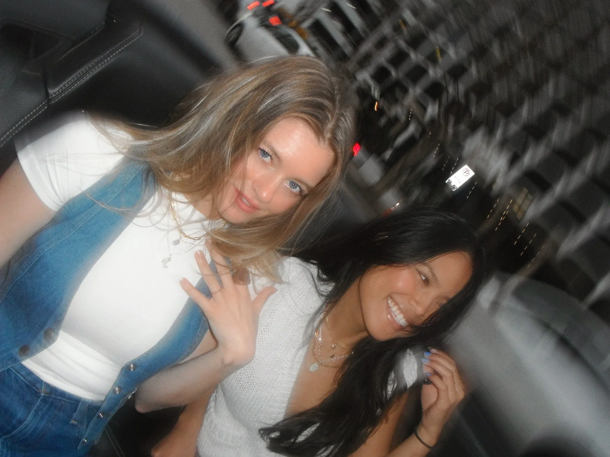 Two young women sitting in a vehicle, smiling. One woman with wavy, light brown hair wearing a white shirt and denim overalls, and the other with long, dark hair and a white top. Background shows a city street with streetlights and buildings.