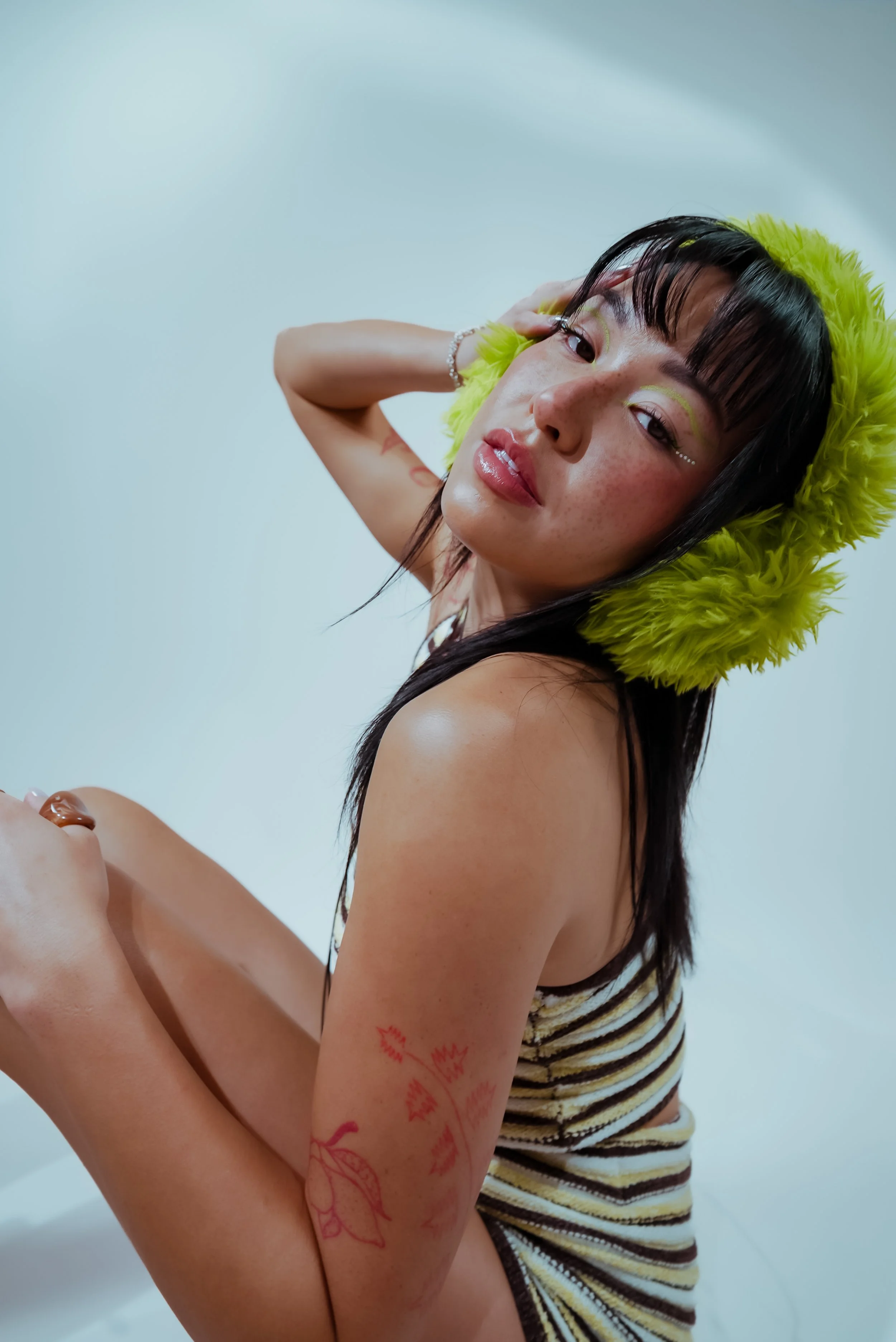 A woman with black hair, wearing a yellow fluffy hat and a striped sleeveless top, poses with her arm raised against a plain white background.