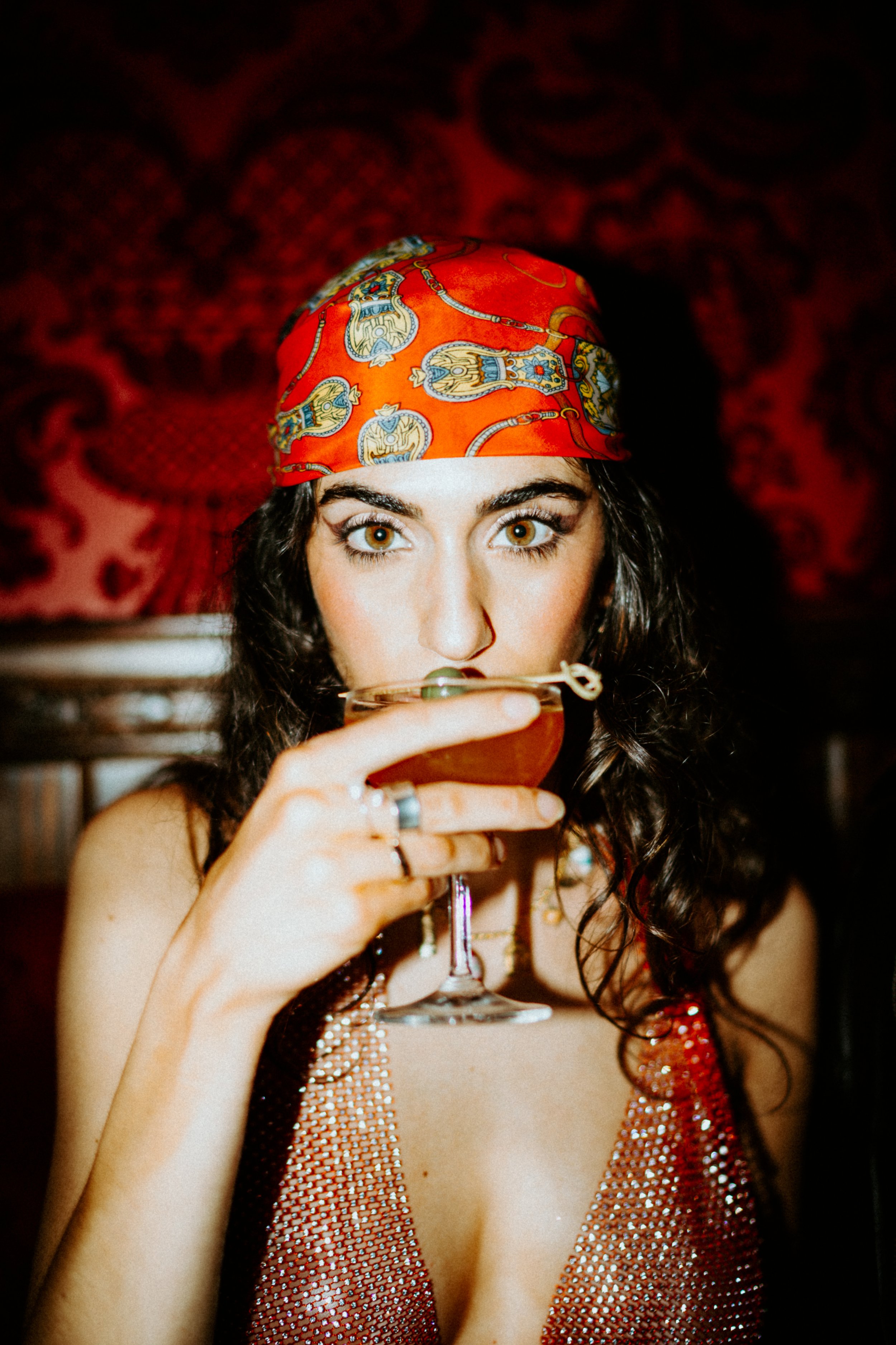 A woman with dark curly hair, wearing a red patterned headscarf, holding a cocktail glass, wearing a sparkly, low-cut top and jewelry.