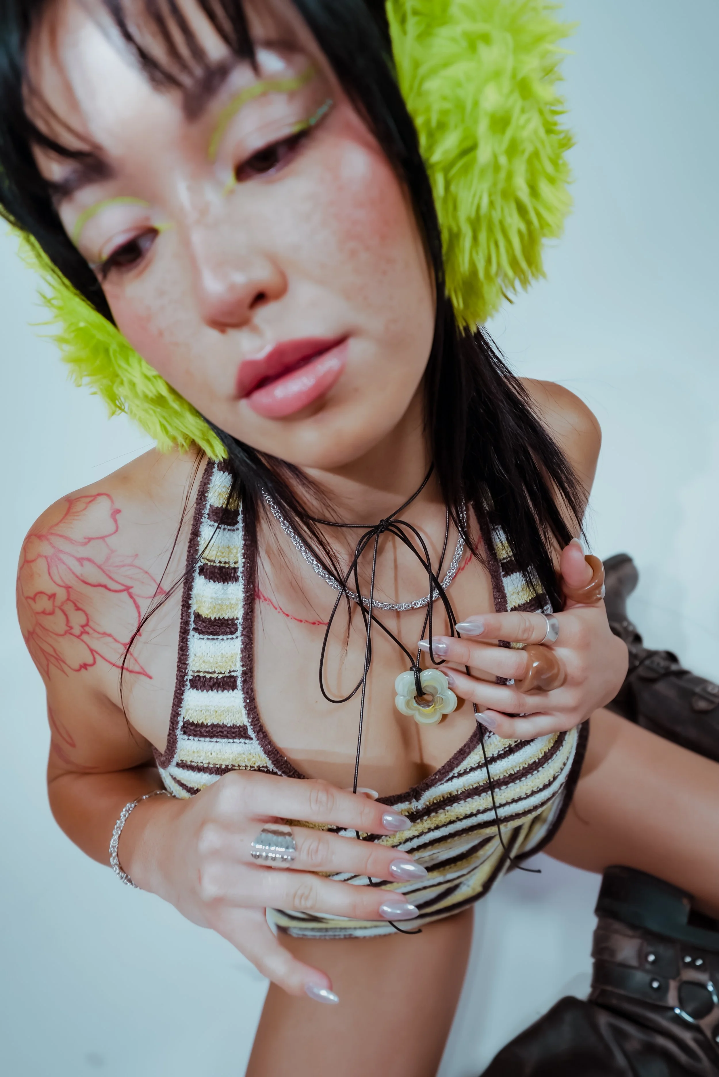 Close-up of a woman with dark hair, wearing fluffy green earmuffs and a striped halter top. She's touching her chest with both hands, displaying necklaces, rings, and bracelets. She has tattoos on her right shoulder and upper arm. The background is p