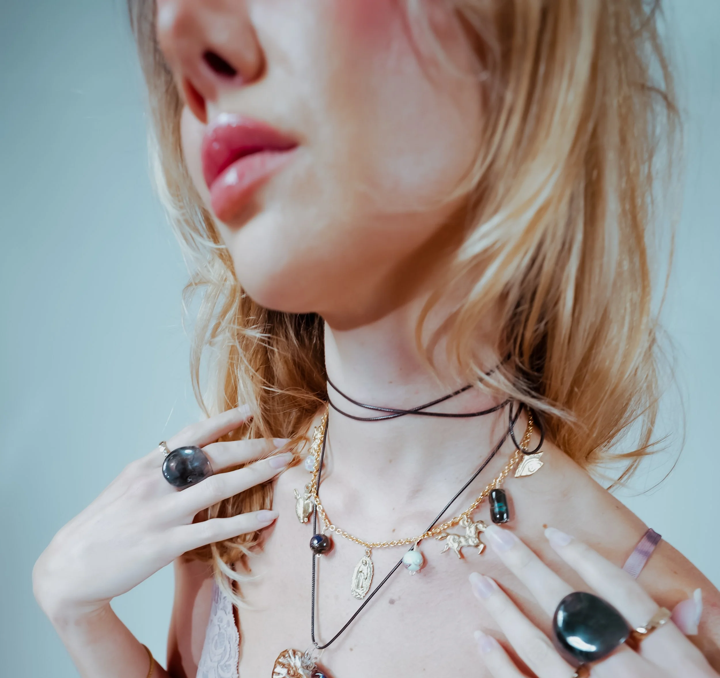 Close-up of a woman's face and chest adorned with various jewelry, including multiple necklaces, rings, and earrings, with her hand touching her chest and a blue background.