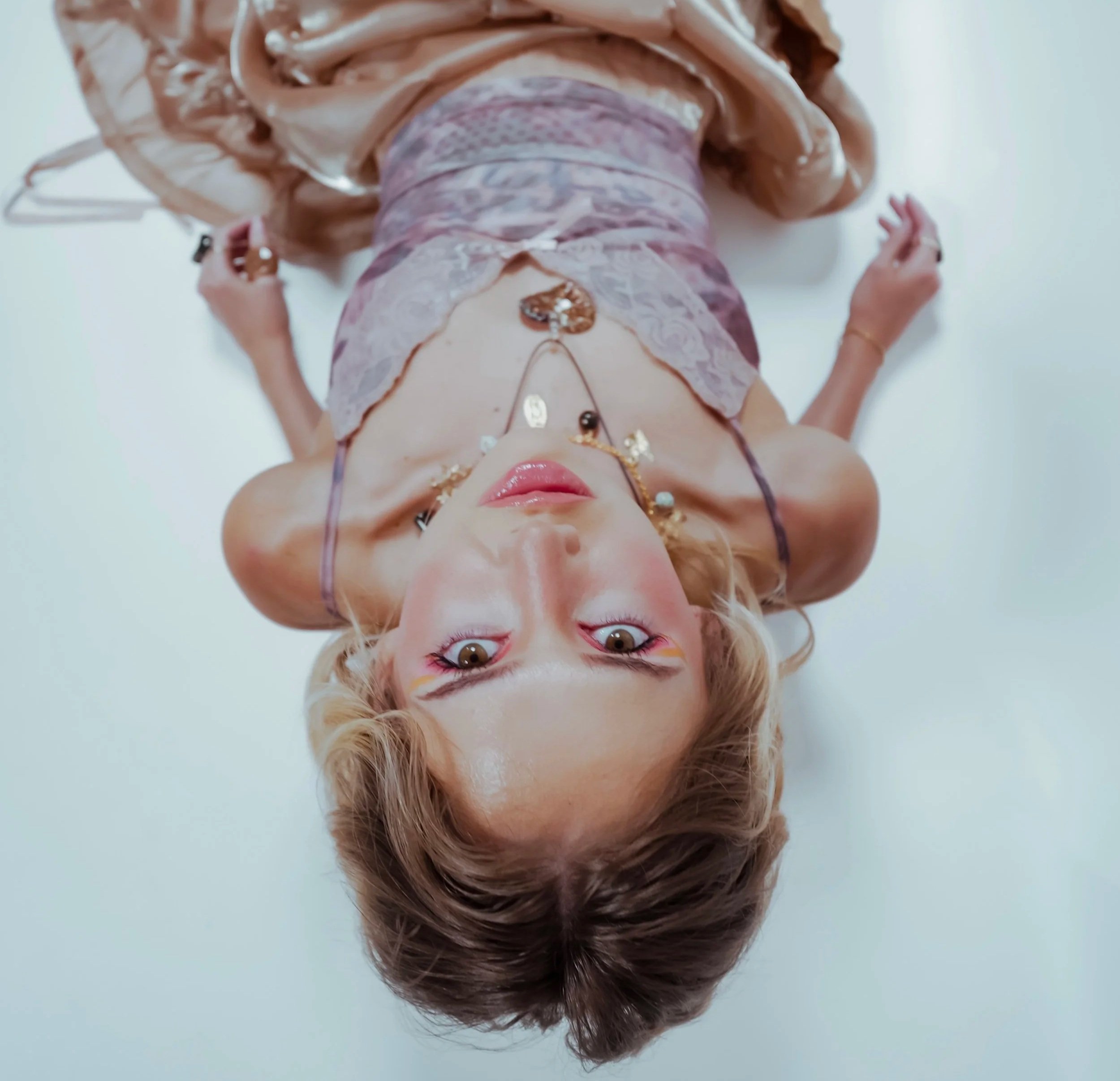 A woman with short blonde hair, wearing multiple necklaces and makeup, lying on her back with her head pointing downward, against a white background.
