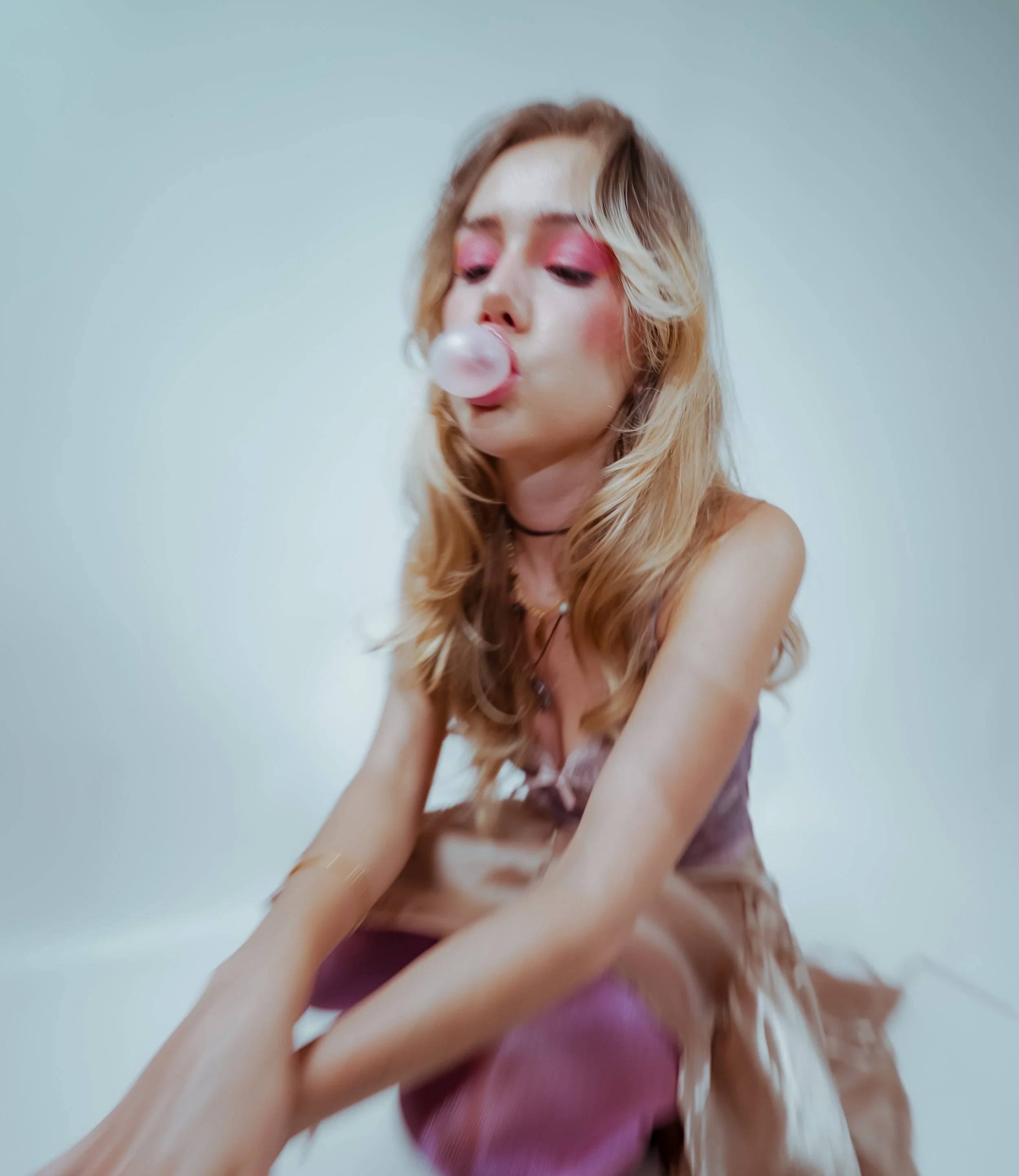 A young woman with long, wavy blonde hair blowing bubbles with bubblegum, wearing pink eyeshadow, a choker, and a purple top, against a plain light-colored background.