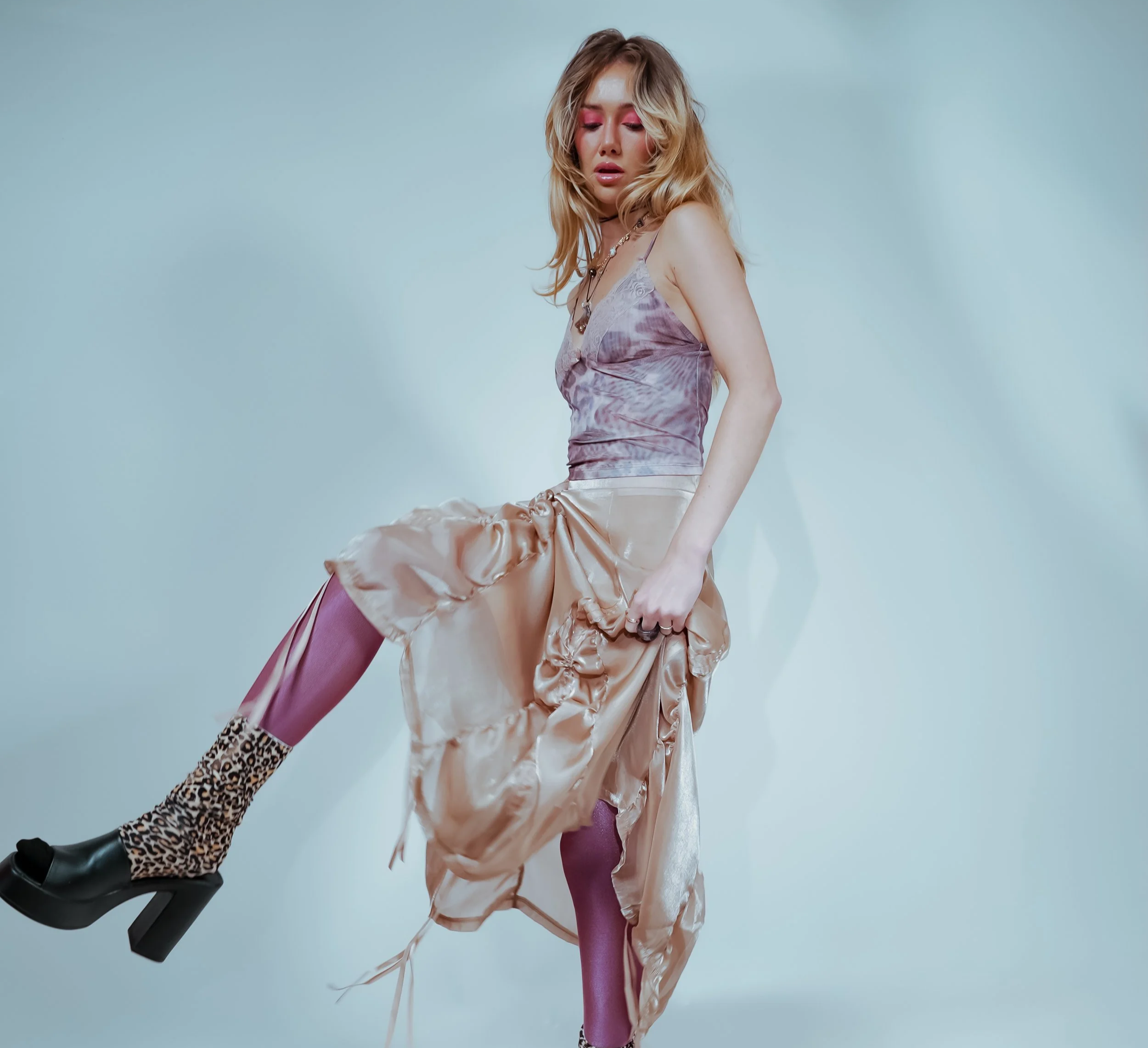 A woman with wavy blonde hair, wearing pink eyeshadow, a silver necklace, a satin dress, pink tights, and leopard print shoes, poses against a plain background.