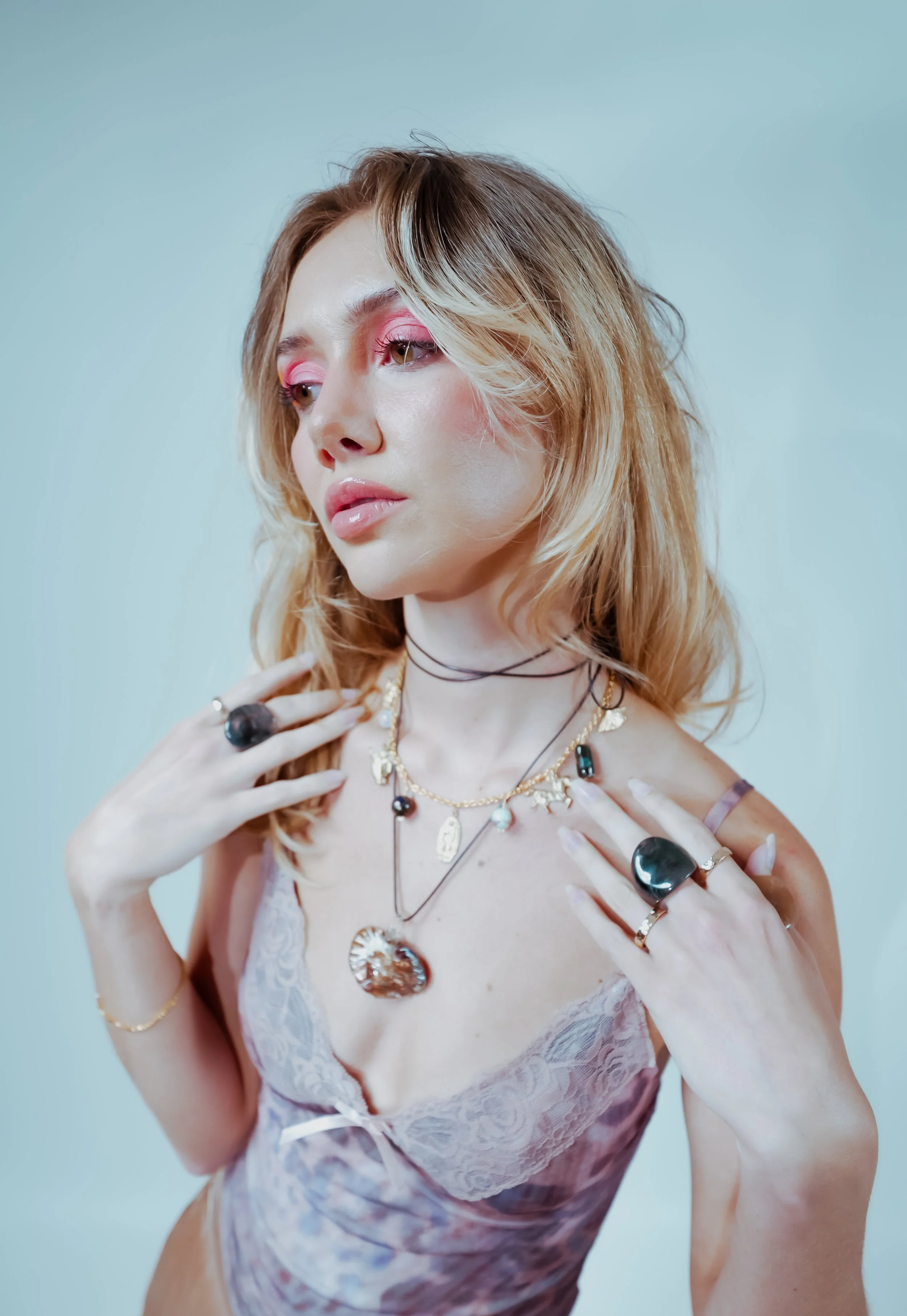 A woman with blonde hair and pink eye makeup wearing layered necklaces and rings, soft pink lipstick, and lace pink top, posing against a light background.
