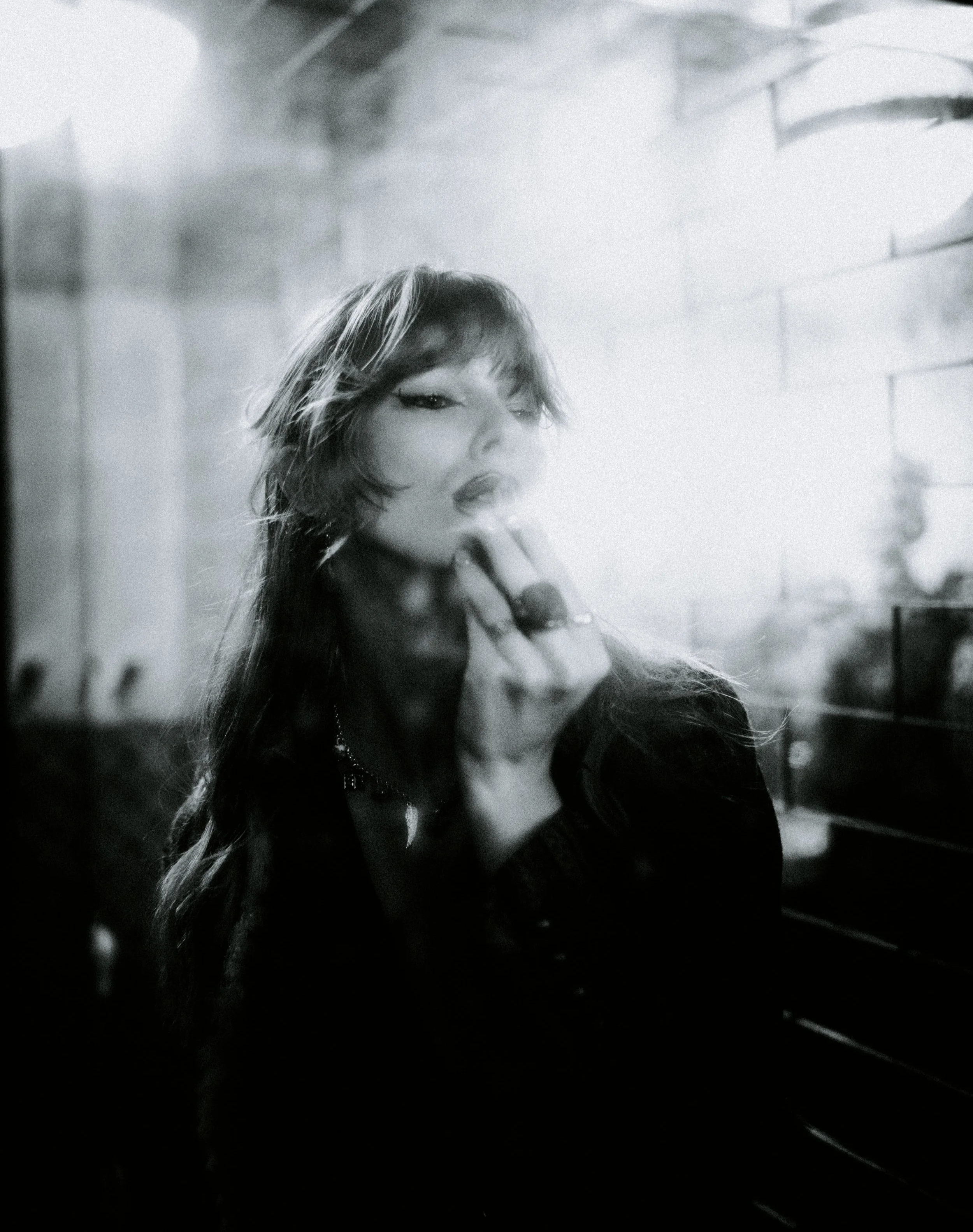 A black-and-white photo of a young woman with long wavy hair, holding a lit cigarette near her lips, with makeup emphasizing her eyes. She appears to be inside a building, with a blurred brick wall in the background.