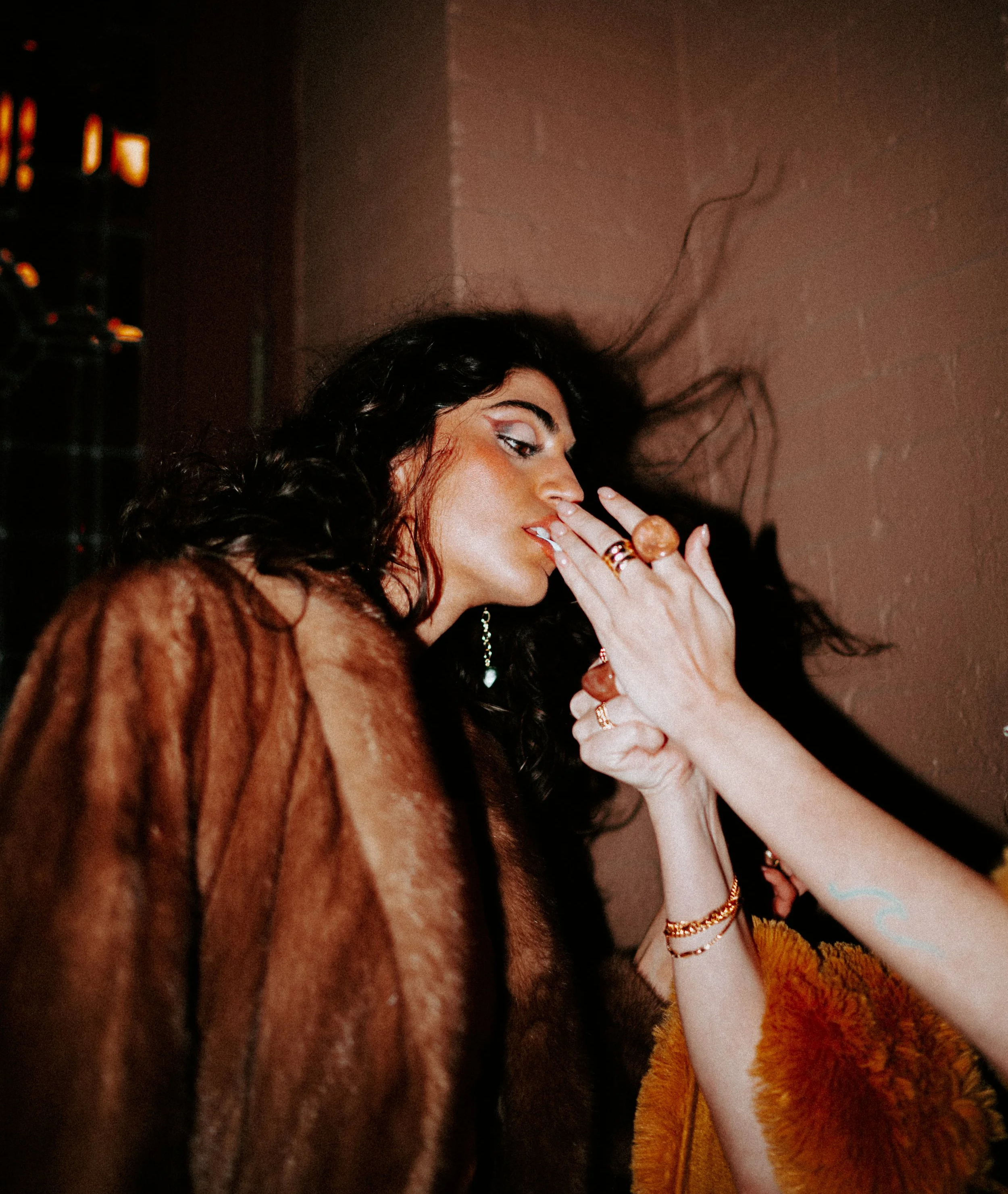 Close-up of a woman with dark curly hair wearing gold jewelry and a yellow fluffy garment, with her hand near her face, touching her lips, against a pinkish background.