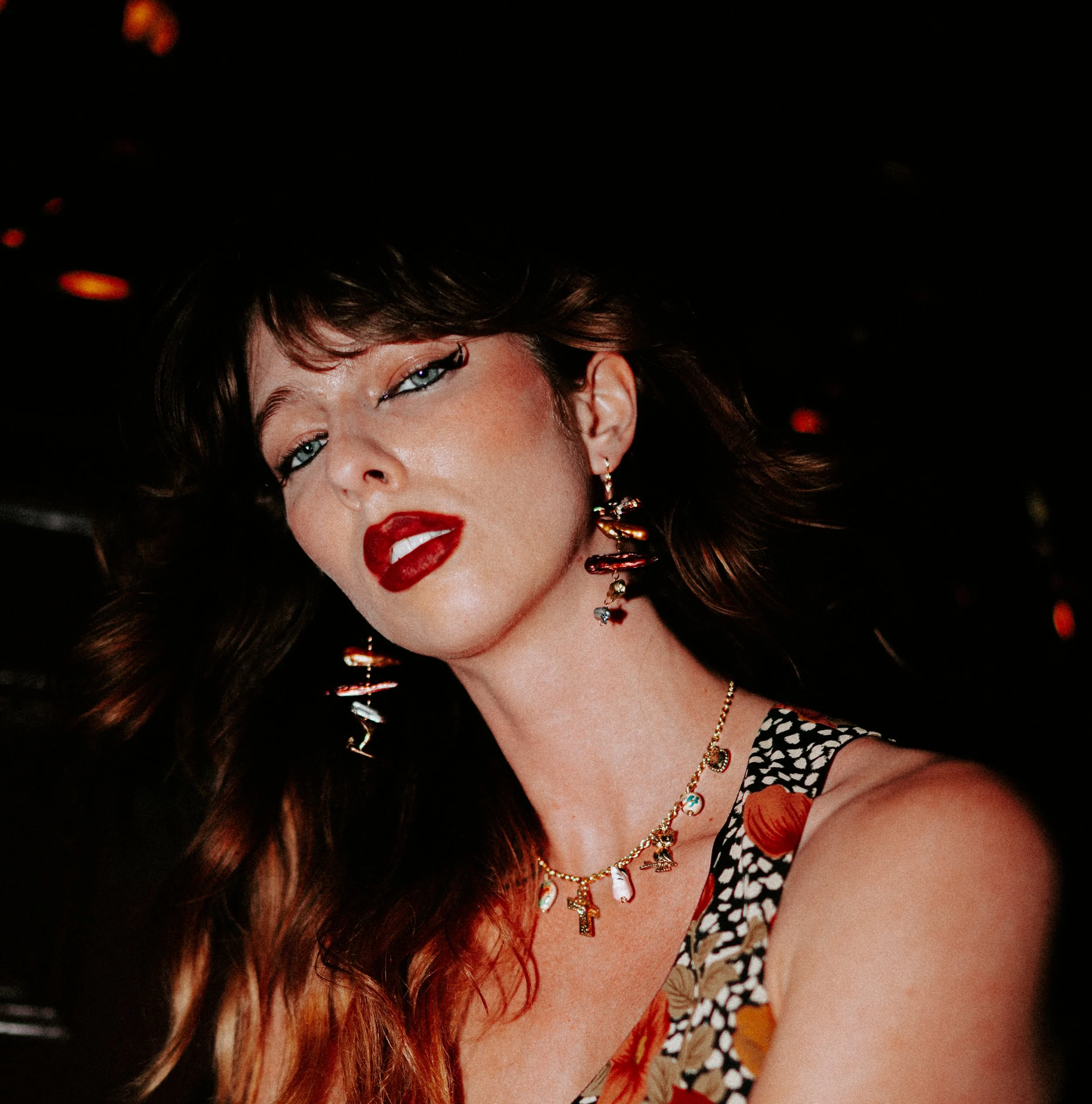 Close-up of a woman's face, showing her eyes, lips, and jewelry, with dark hair and a patterned outfit, against a dark background with orange bokeh lights.