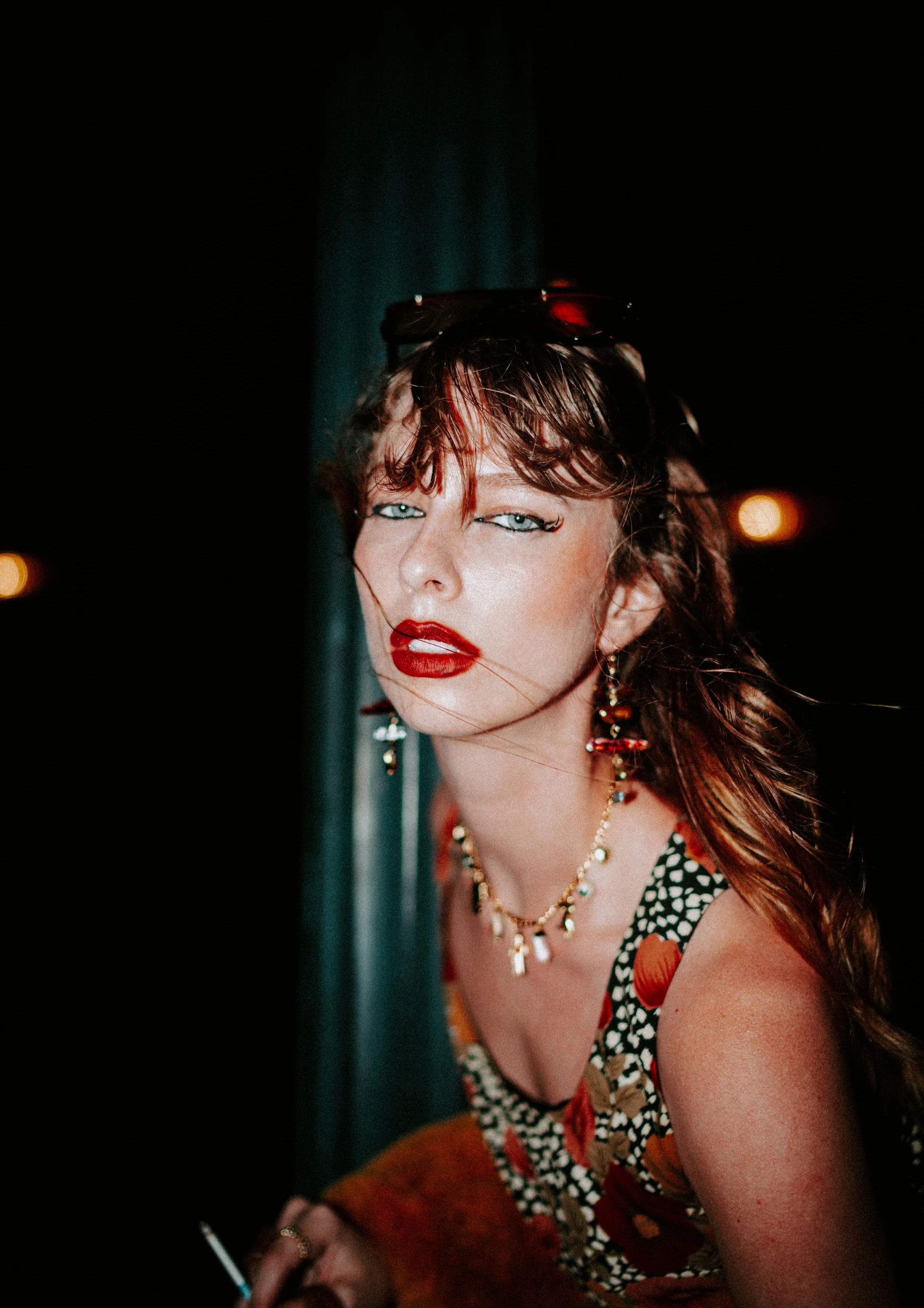 A woman with red hair and blue eyes wearing bold makeup, colorful earrings, a patterned dress, and holding a cigarette, is seen in a dark setting with blurred lights in the background.