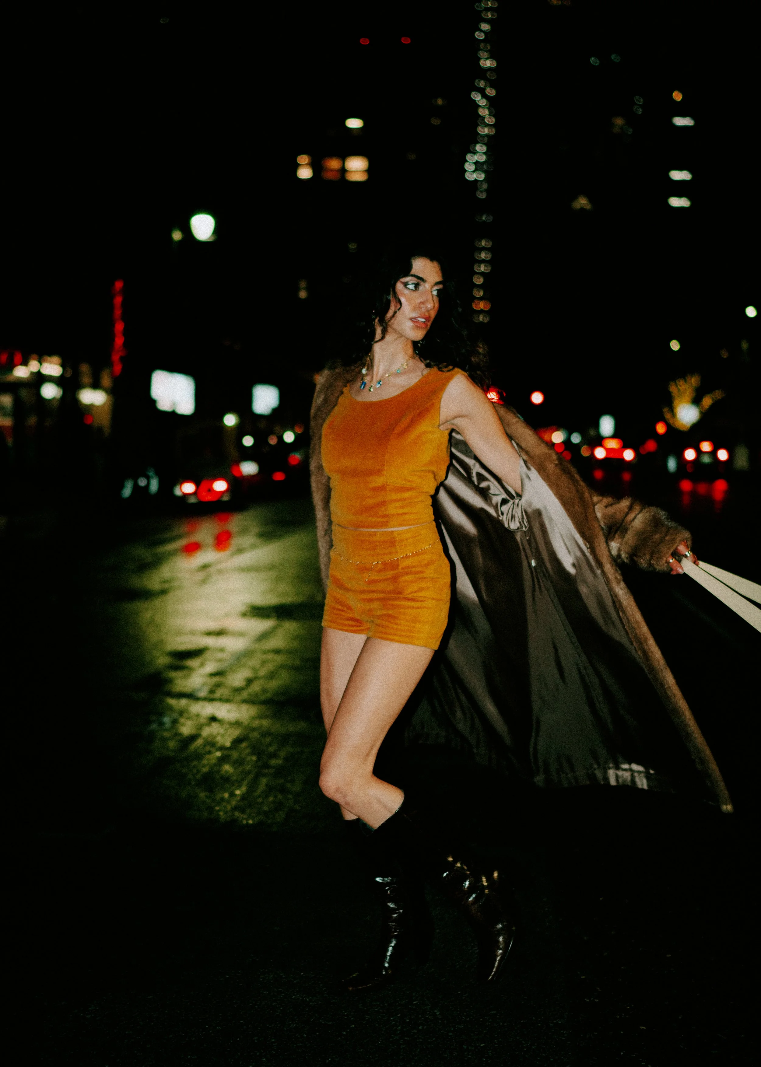 A woman with dark curly hair wearing an orange dress and holding a fur coat on her shoulder stands on a city street at night with blurred city lights and buildings in the background.