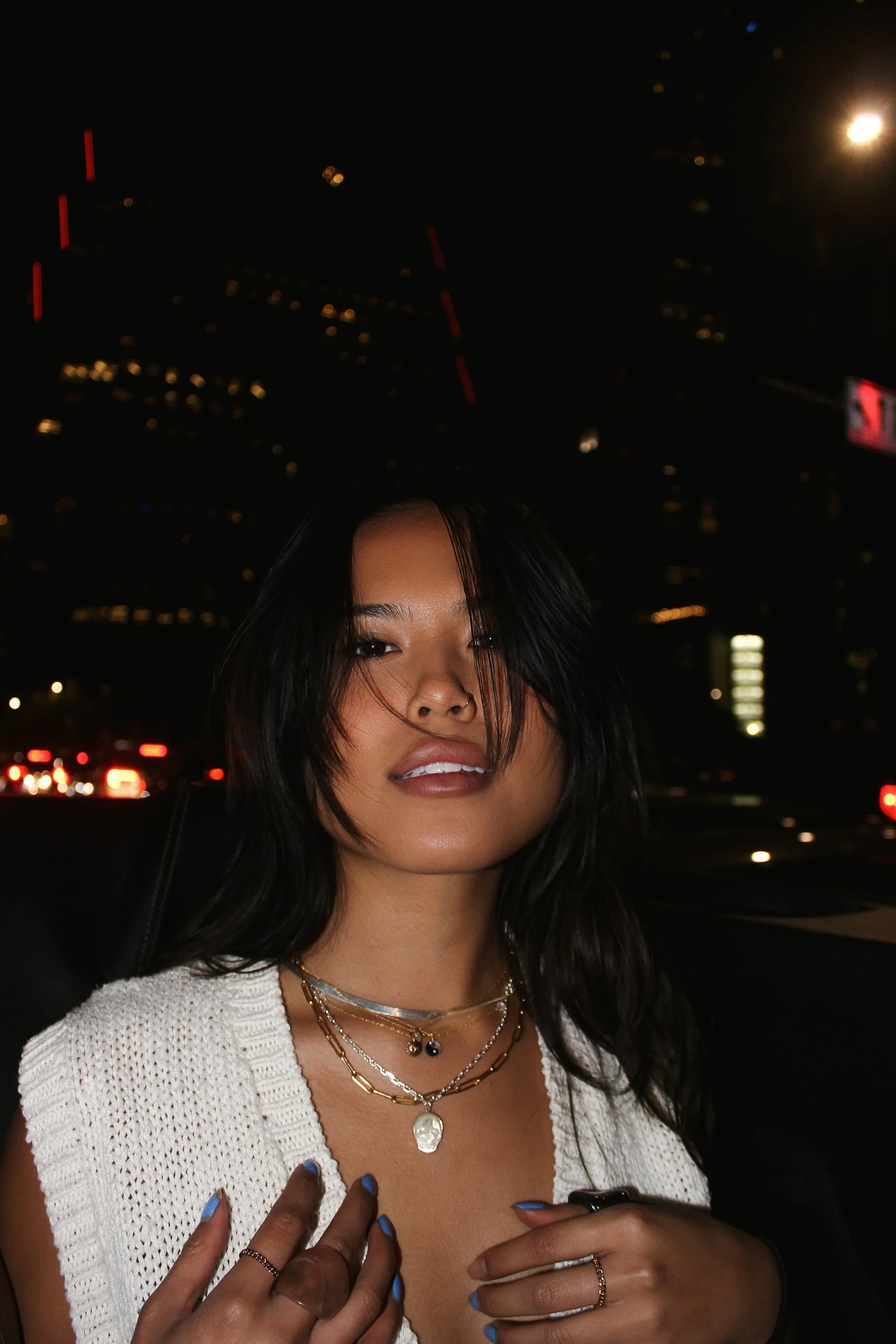 A young woman with dark hair smiling at night, wearing layered necklaces, a white knit top, and blue nail polish. Her face is partially obscured by her hair, and there are city lights and buildings in the background.