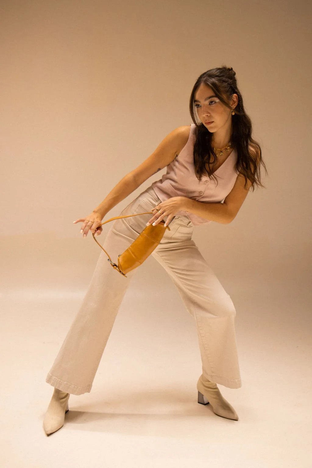 A woman dressed in beige pants and a pink sleeveless top standing against a plain beige background. She is holding a small orange purse and striking a stylish pose.