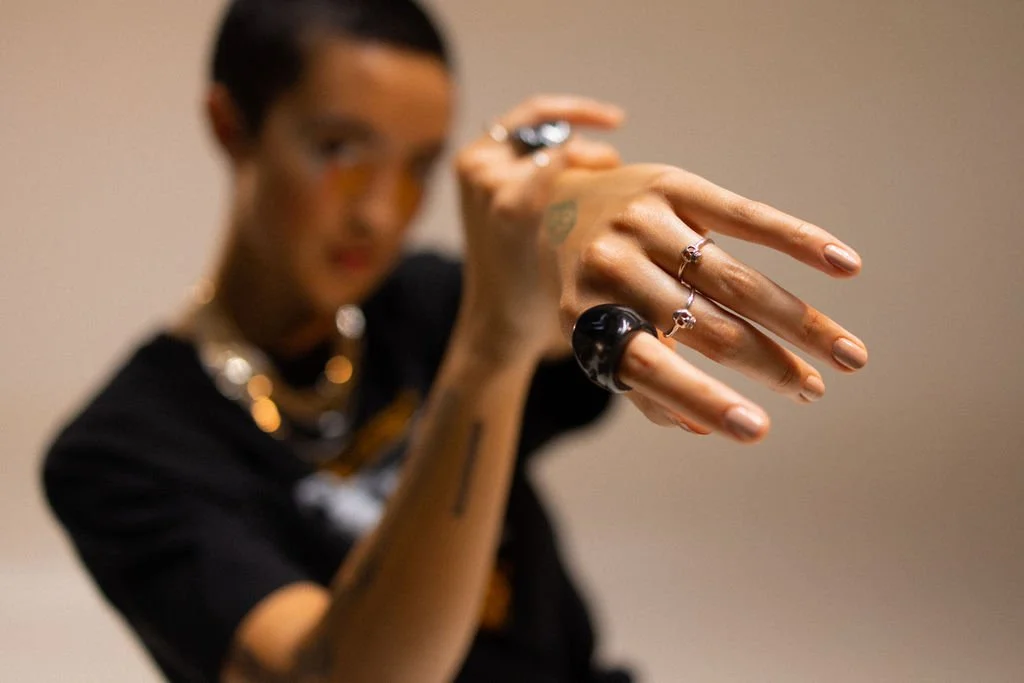Person with tattoos and jewelry showing their hand with rings in focus, face blurred in background.
