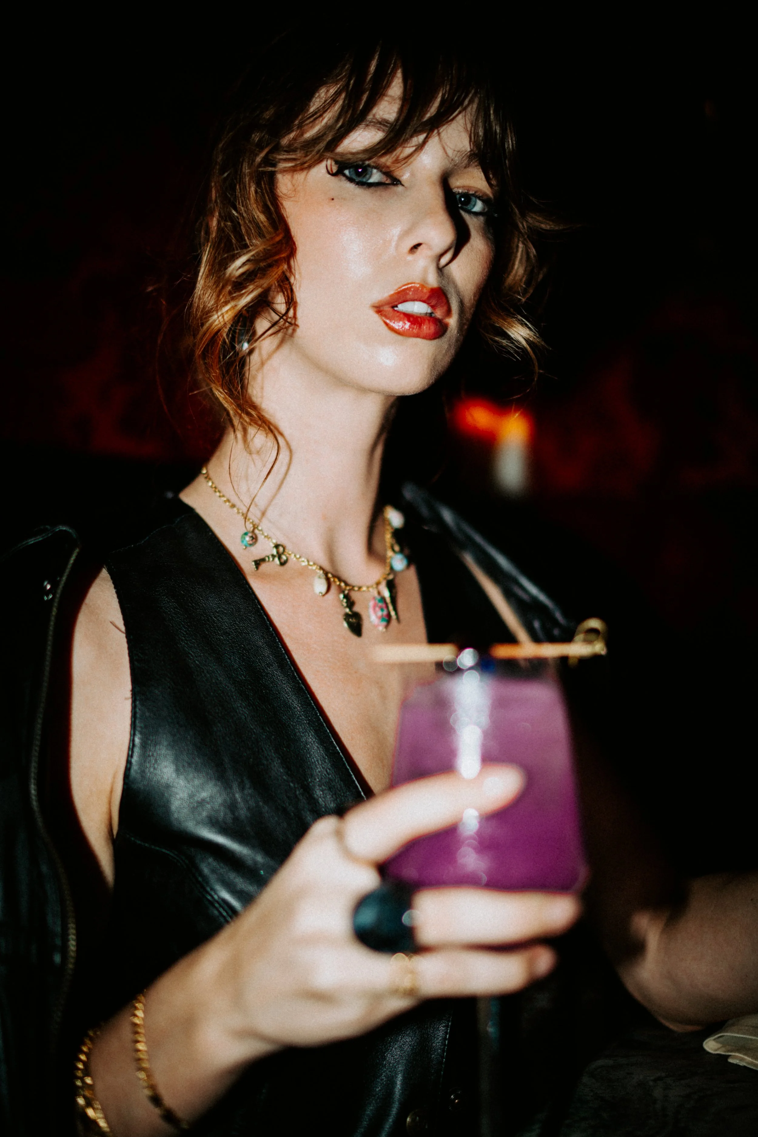 A woman with wavy brown hair, wearing makeup, jewelry, and a black leather vest, holding a purple phone and looking at the camera.
