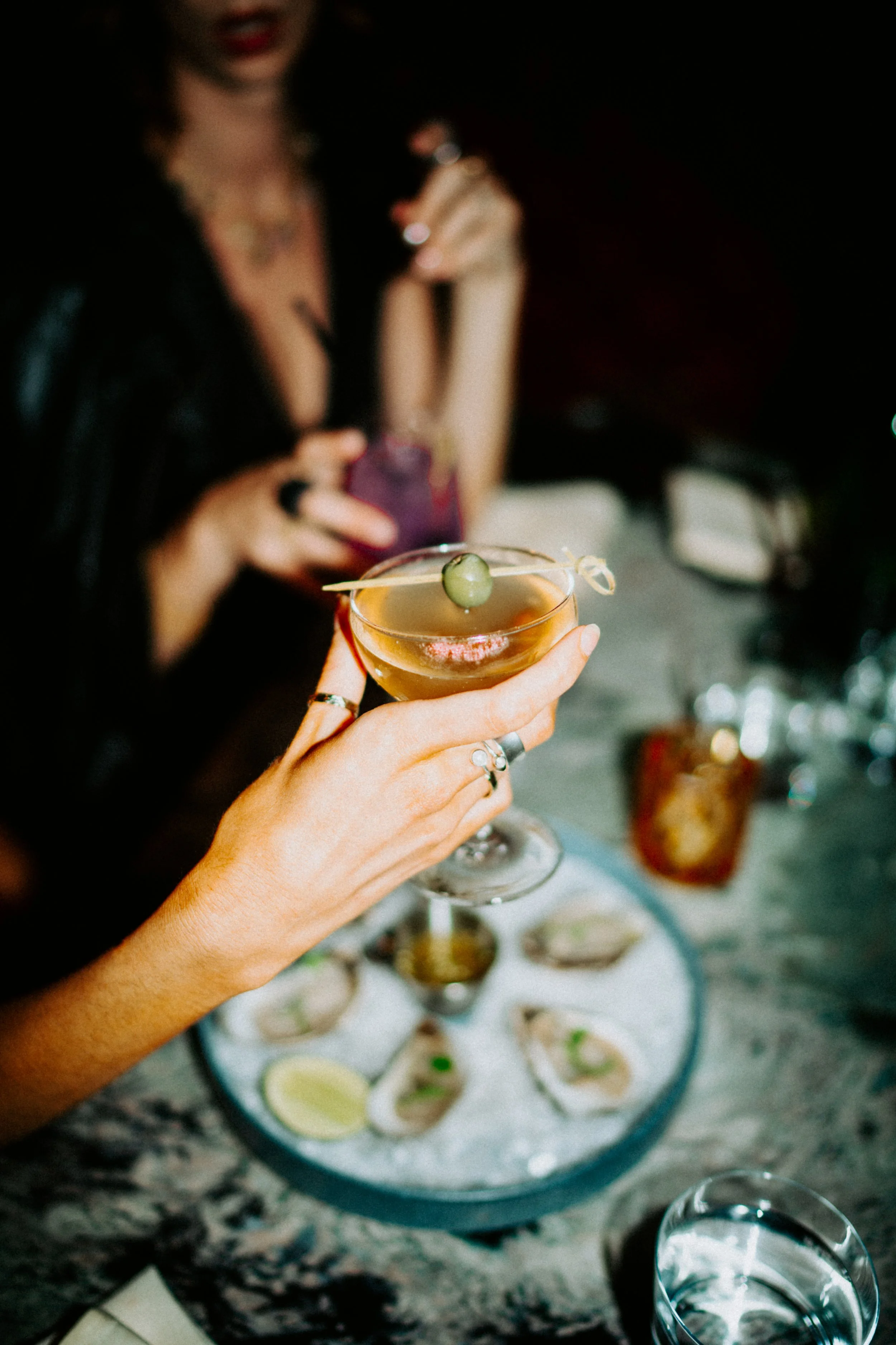 Close-up of a person holding a cocktail glass with an olive garnish, with a woman in the background preparing drinks at a bar or party setting.