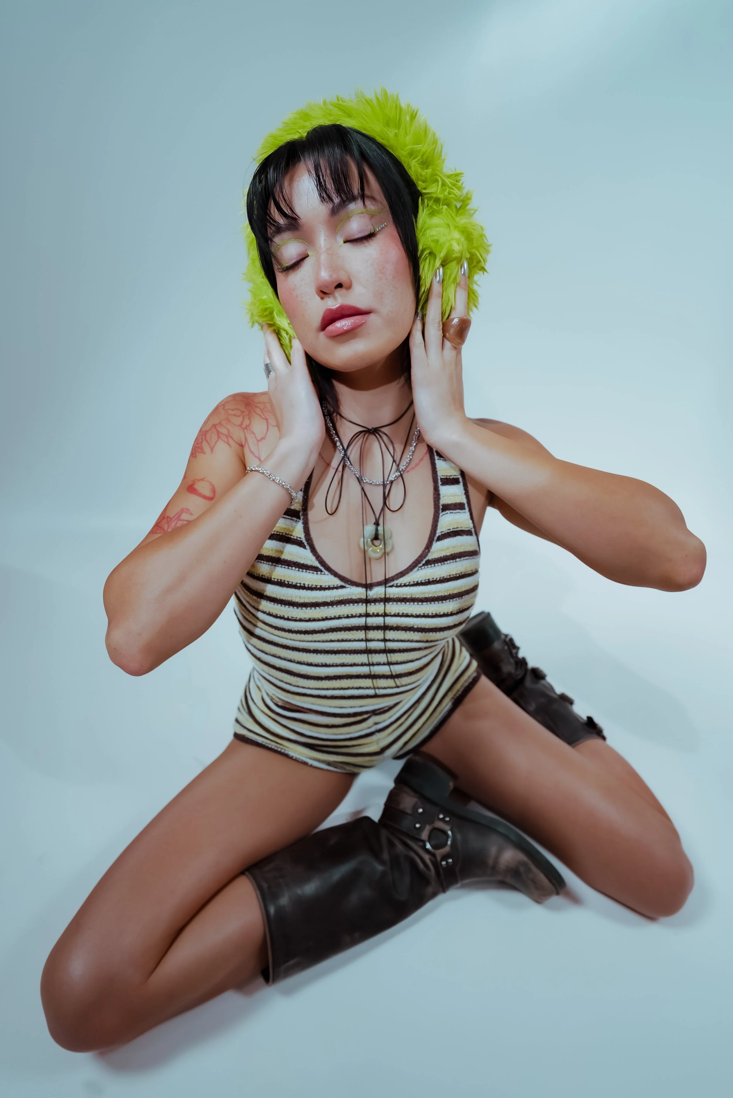 A woman with black hair and tattoos, wearing a striped tank top, earrings, and layered necklaces, posing with her eyes closed. She is touching her head with both hands, and is seated with her legs apart on a white surface. She is wearing black boots.