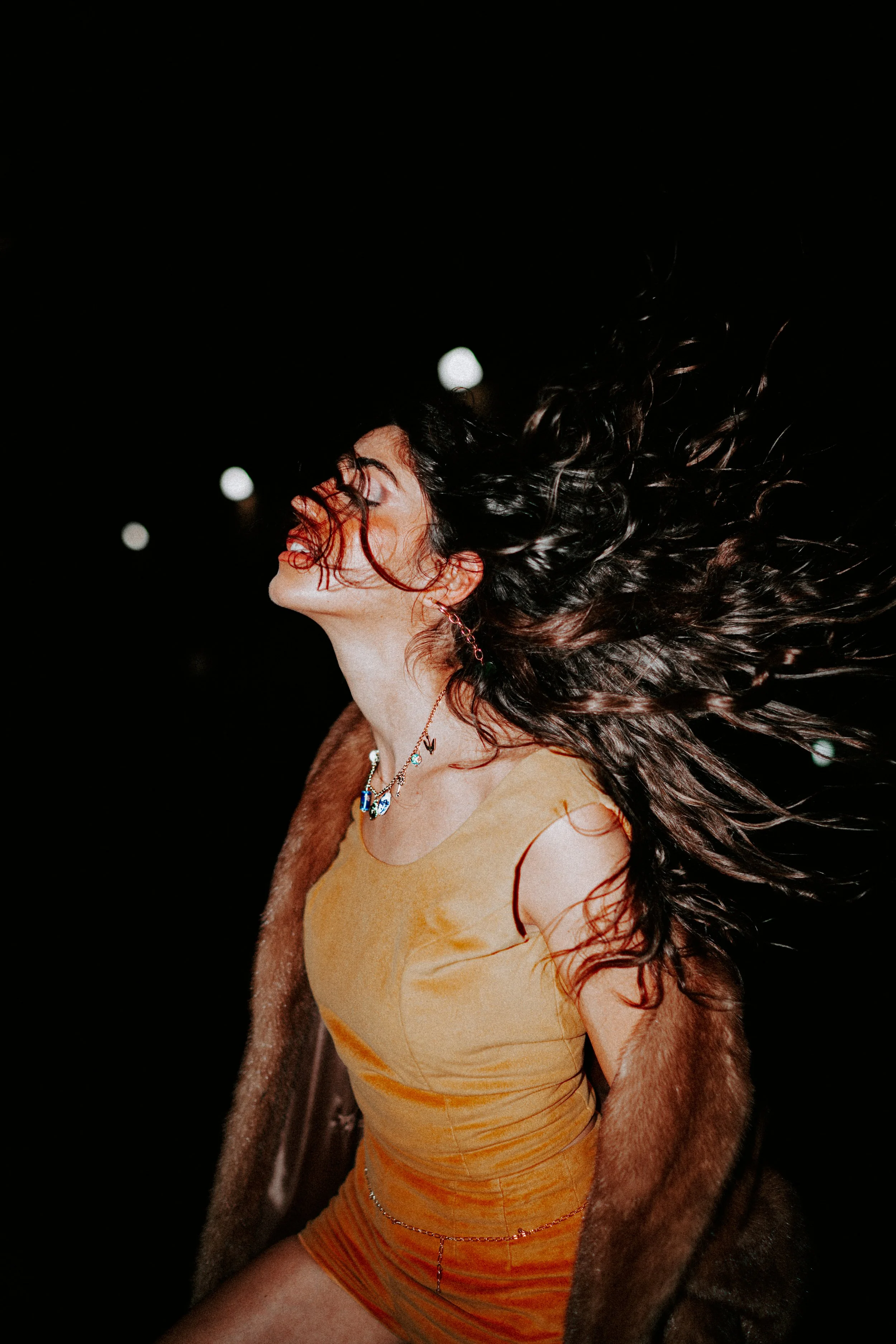 A woman with long, curly dark hair is captured mid-motion against a black background. She is wearing a beige dress, a fur coat draped over her shoulders, and jewelry including earrings, a necklace, and a waist chain.