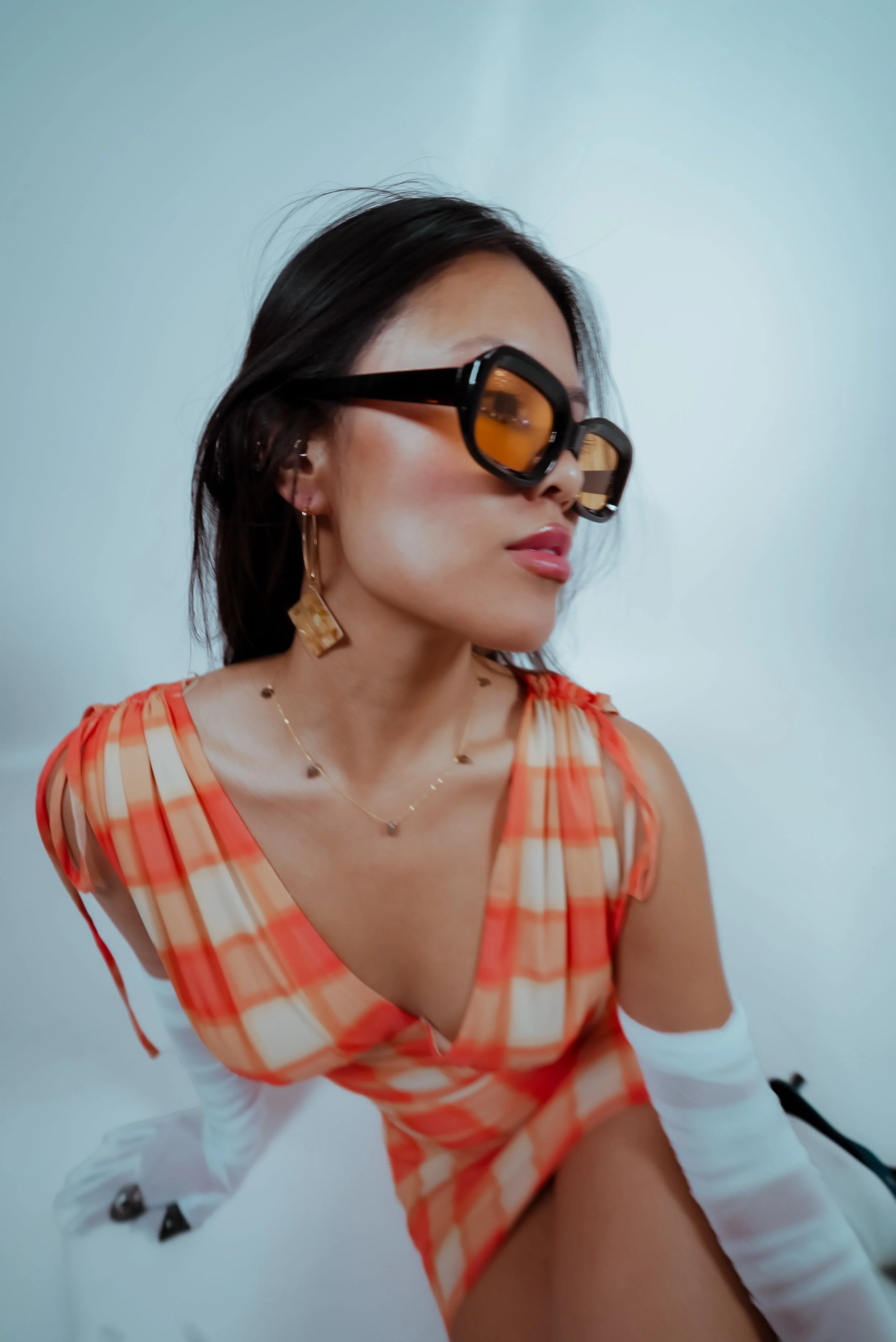 A woman with dark hair wearing large black sunglasses with amber lenses, a gold necklace, hoop earrings, and a coral and white checkered dress with white arm coverings. She is in a close-up pose against a plain white background, with her face turned sideways.