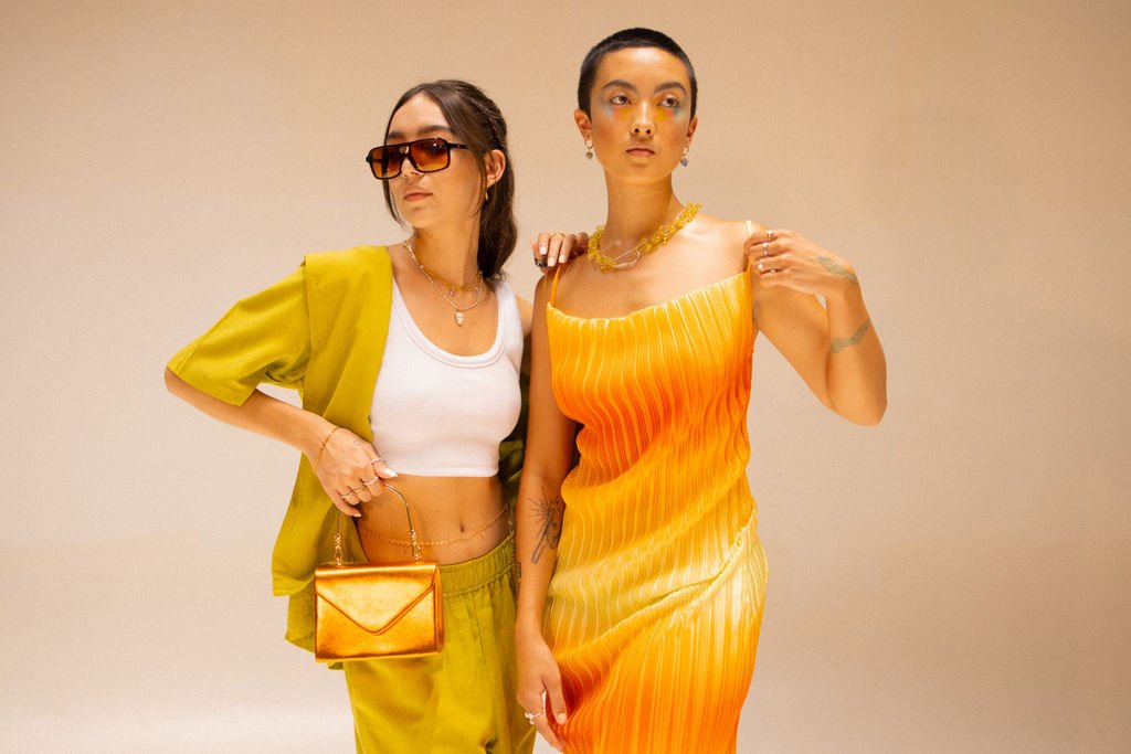 Two women posing together indoors against a beige background. The woman on the left wears sunglasses, a white cropped top, and a green jacket with matching pants, carrying a small gold purse. The woman on the right wears a sleeveless orange and yello