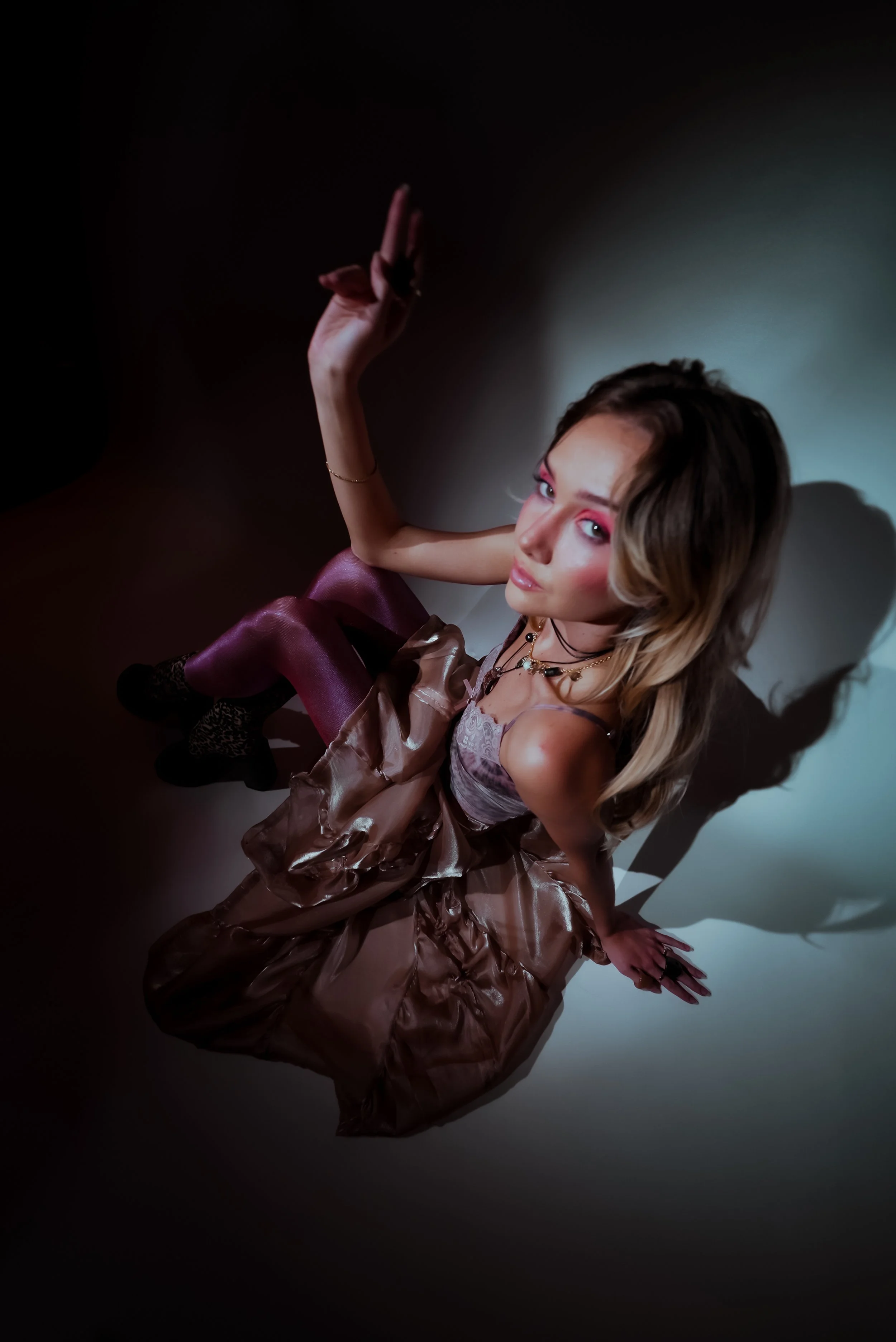 A woman with long, wavy blonde hair, pink eye makeup, and jewelry, sitting on the floor against a plain background, wearing a shiny bronze dress with puffed sleeves and pink tights, looking up at the camera.