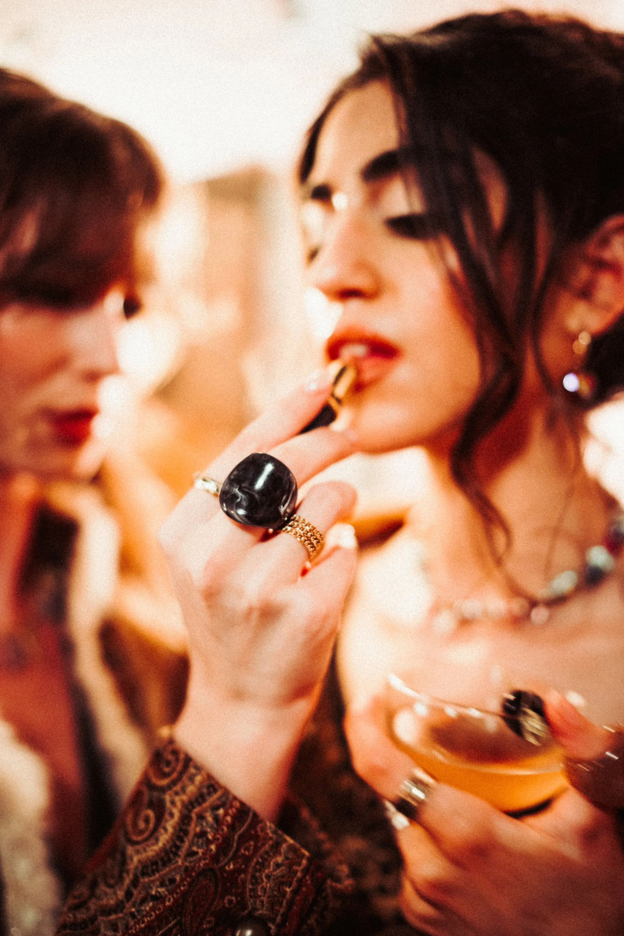 Two women at a social gathering, one applying lipstick and the other holding a glass of drink.
