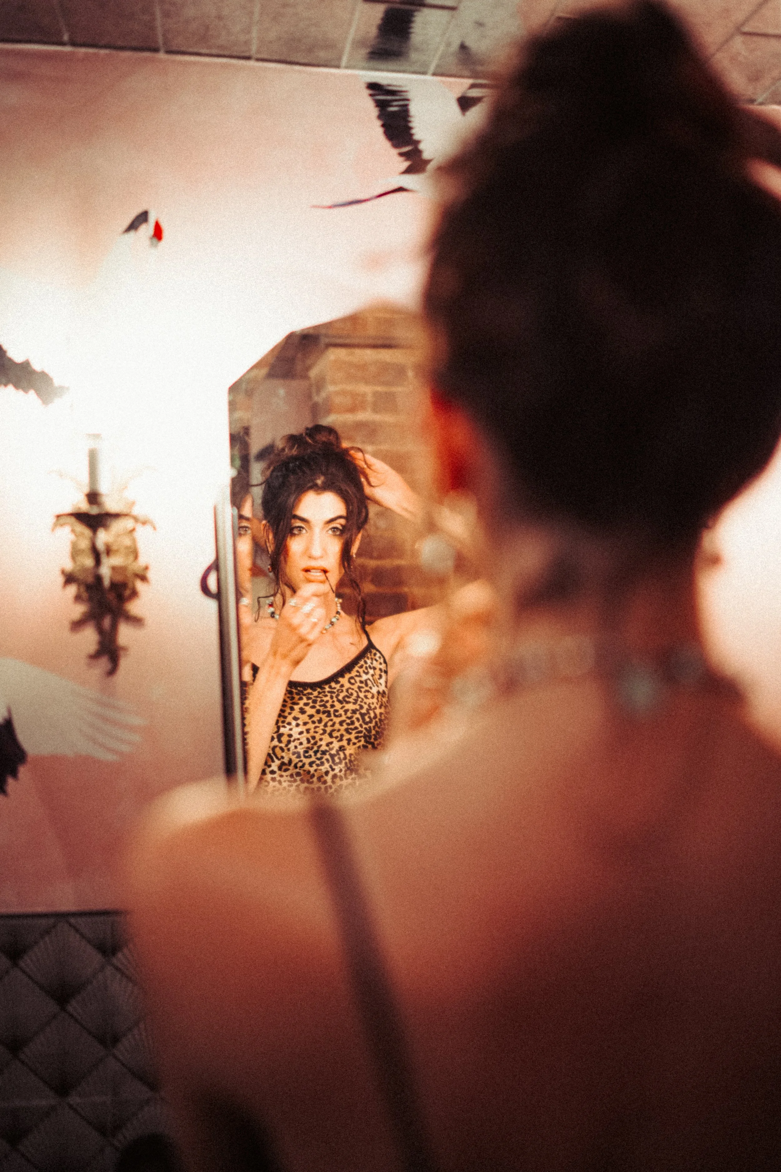 A woman taking a mirror selfie in a leopard print top, with dark curly hair and makeup, in a room with brick wall and Halloween decorations.