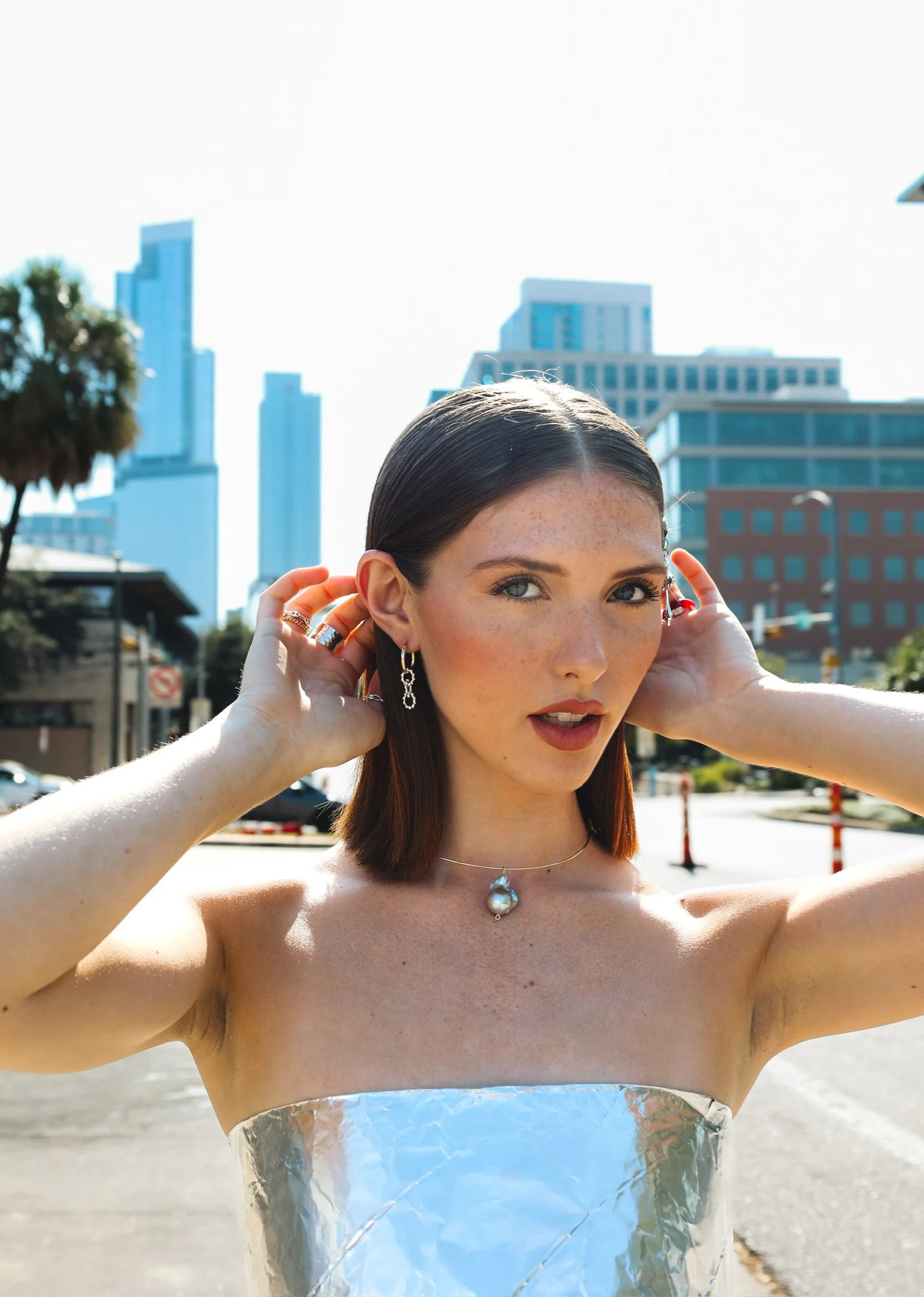 A woman with straight brown hair and blue eyes adjusting her earrings, standing outdoors with tall buildings behind her, wearing jewelry and a silver foil dress, with sunlight creating bright highlights and shadows.